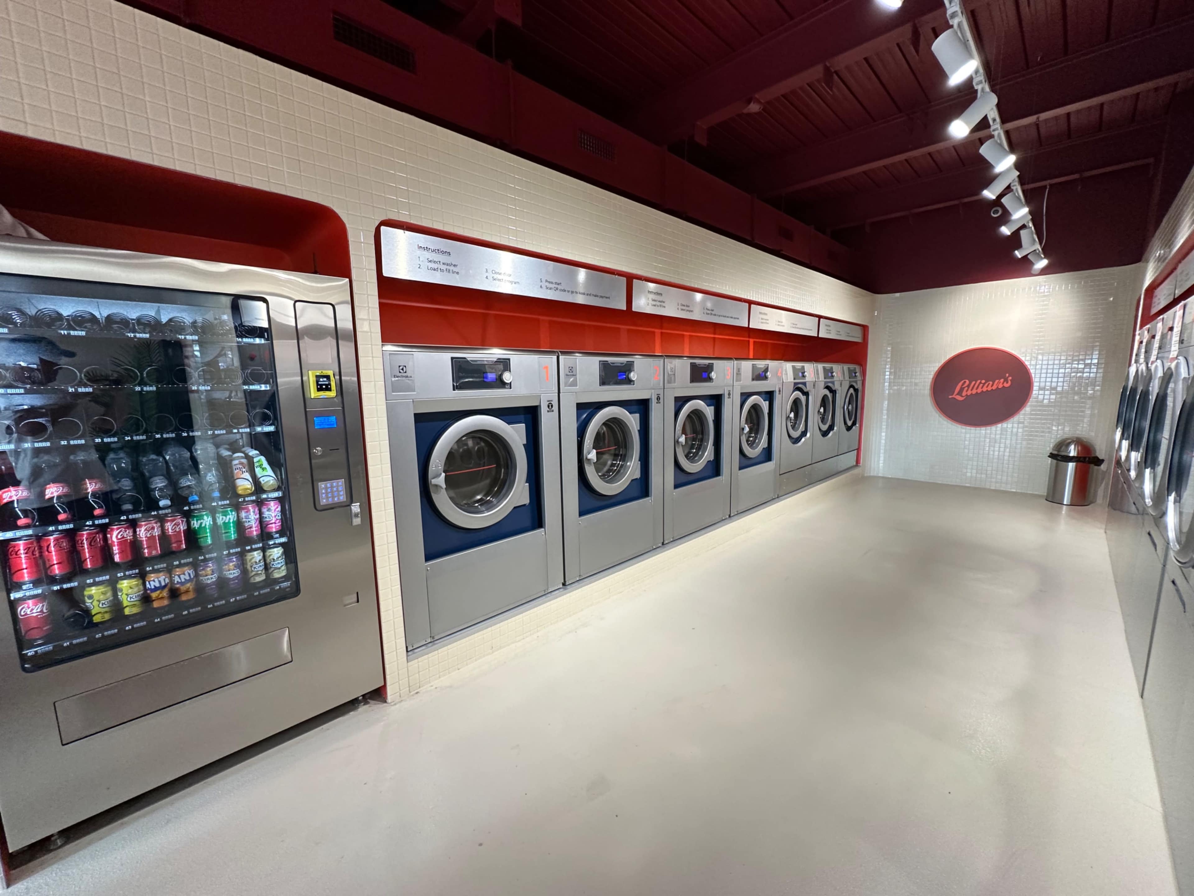 The image shows a modern laundromat with several washing machines lined up against a brightly lit wall, accompanied by a vending machine on the left.