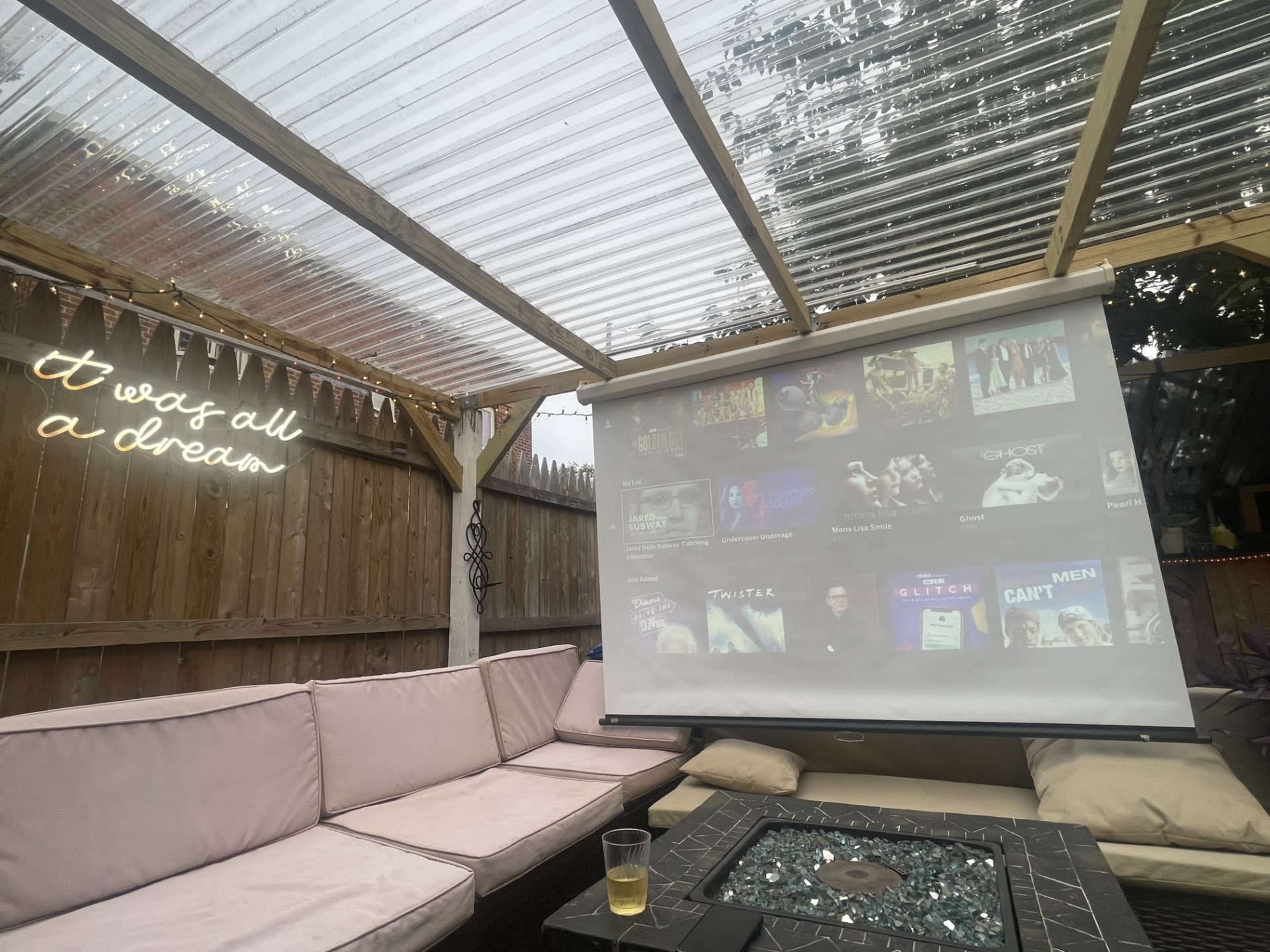 A cozy outdoor seating area features a projector screen displaying a selection of movies, with a drink placed on a table nearby.