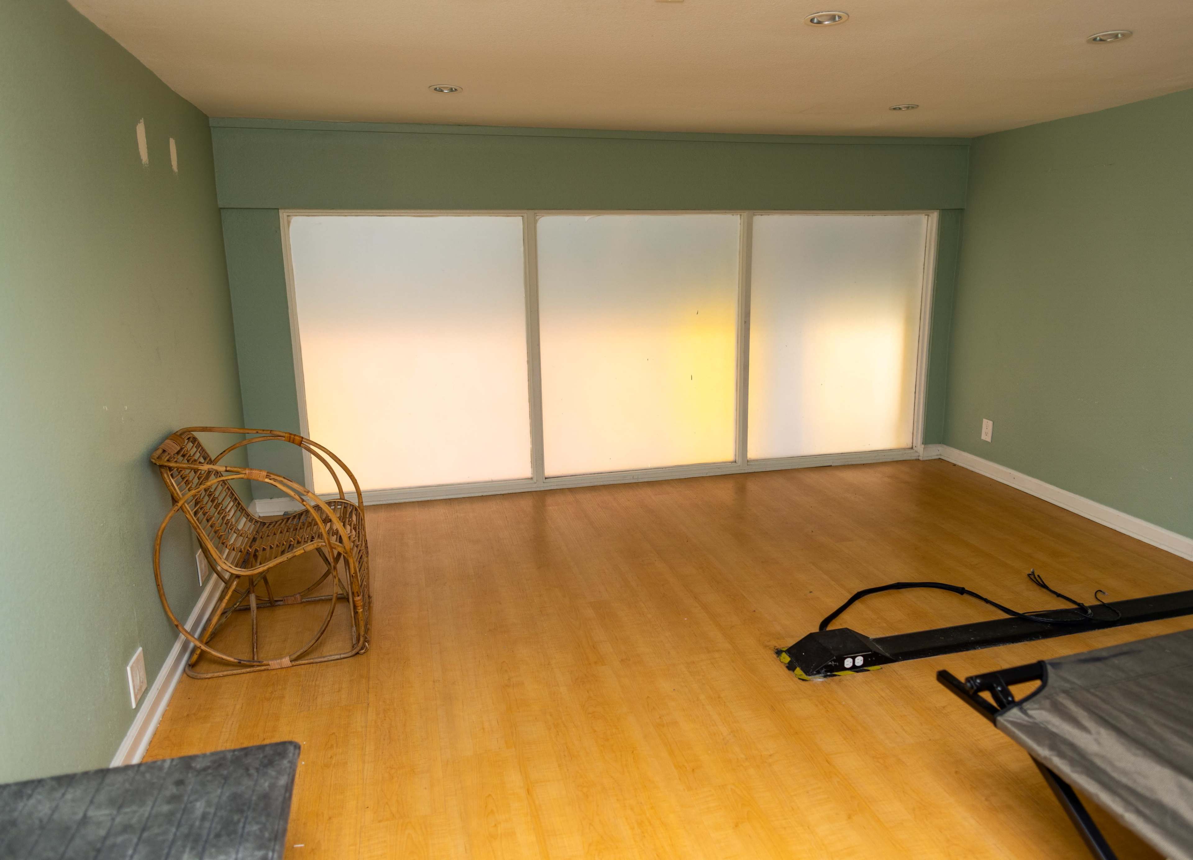 The image shows an empty room with a light green wall, a wooden floor, a rattan chair in the corner, and large frosted windows.