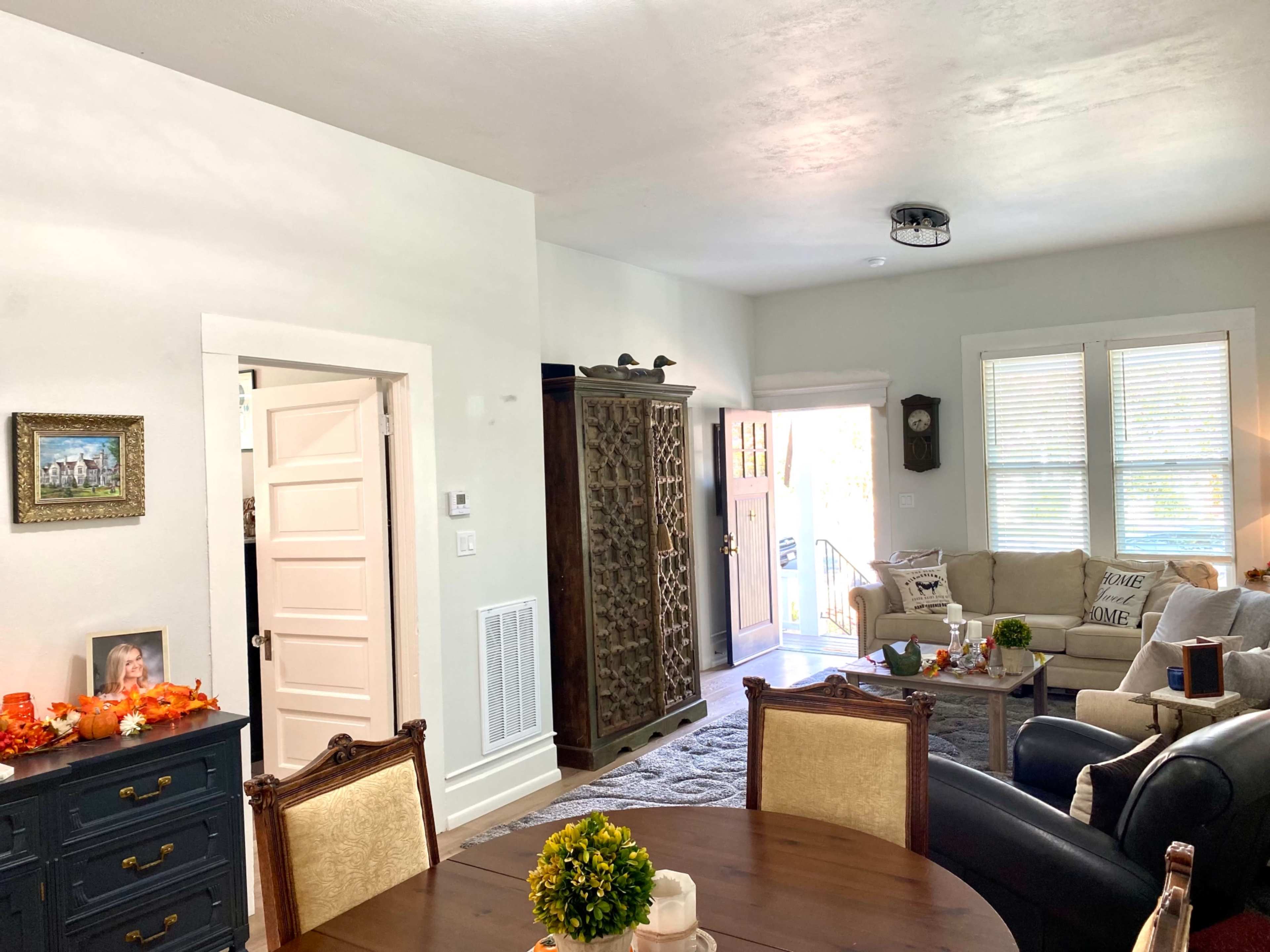 A cozy living space with a dining area featuring a round table and chairs, a sofa set, and a textured rug, alongside a decorative cabinet and bright natural light from the doorway.