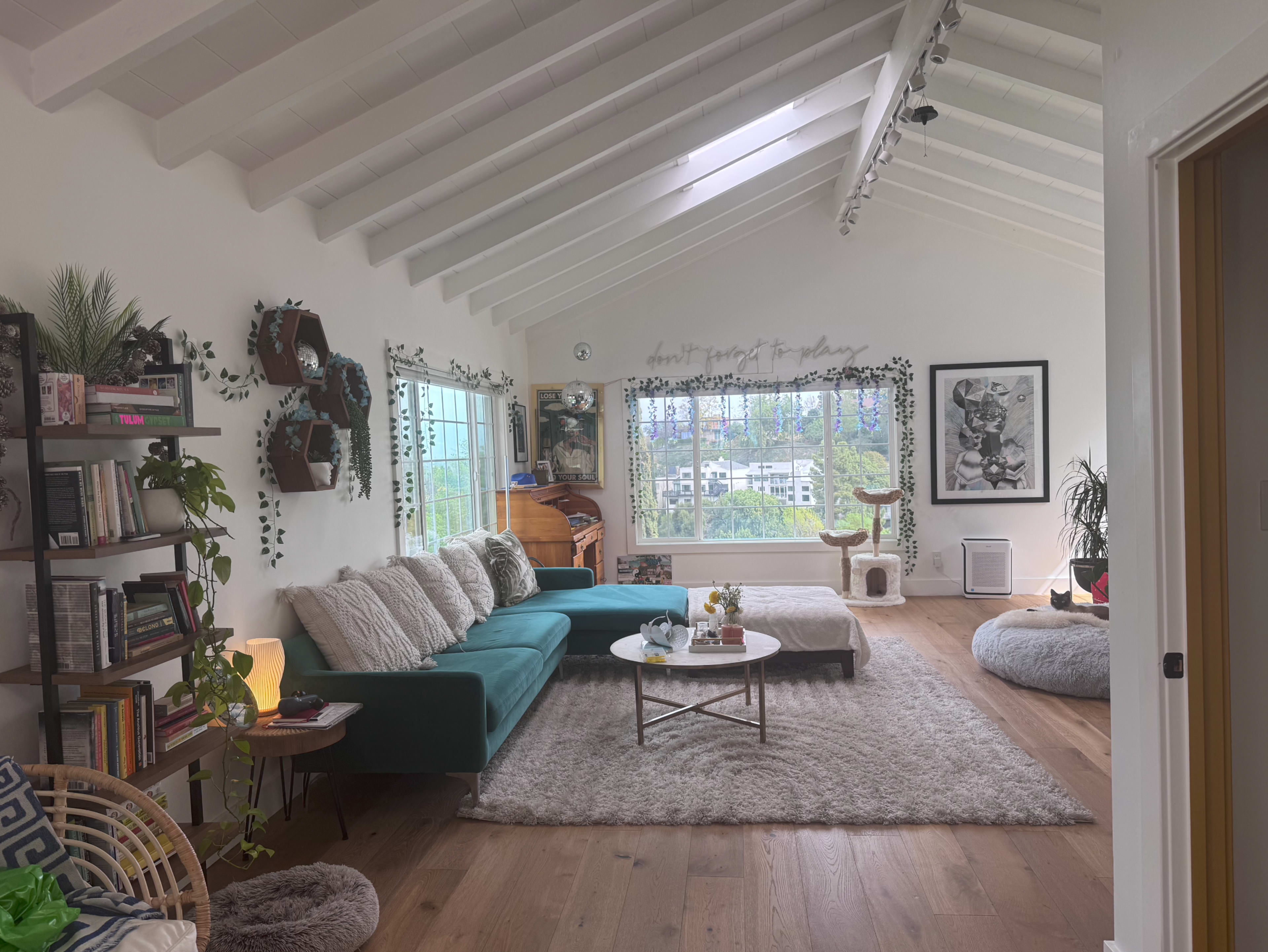 The living room features a teal couch, a large rug, and panoramic windows with greenery outside, along with shelves filled with books and decorative plants.