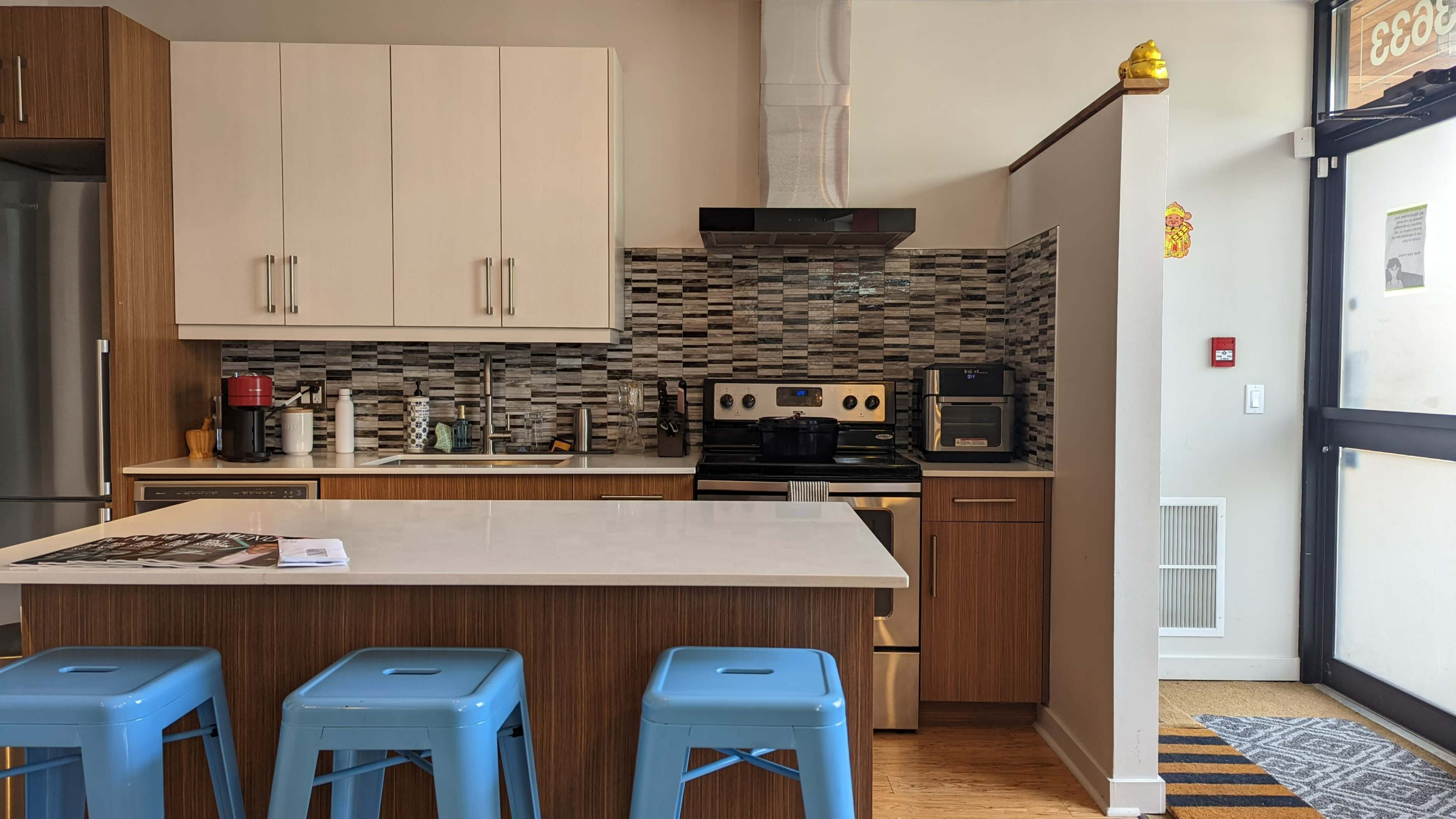 The image shows a modern kitchen with a central island, blue stools, stainless steel appliances, and a tiled backsplash.
