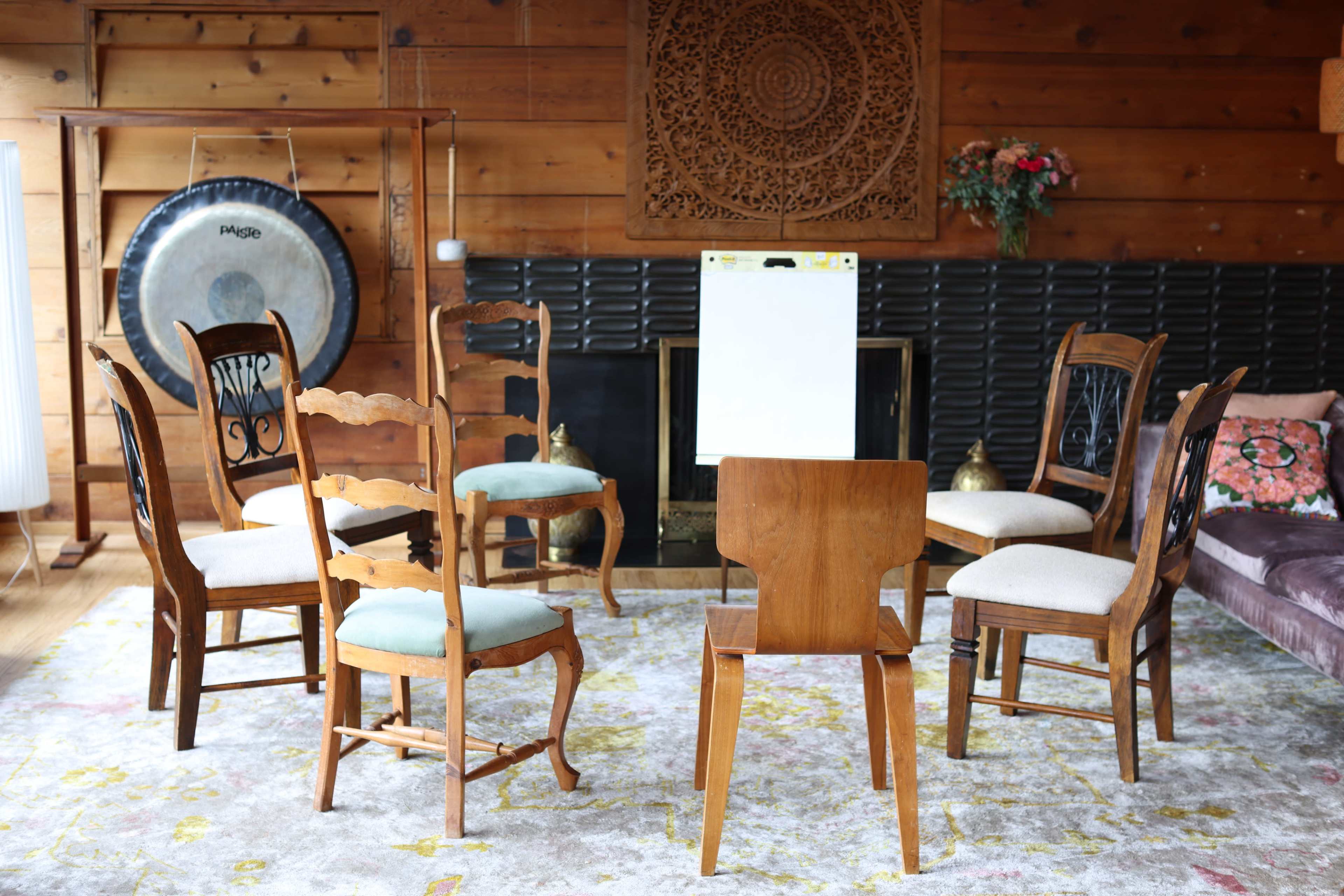 A circle of six wooden chairs surrounds a blank easel in a room with wooden walls and a patterned rug.