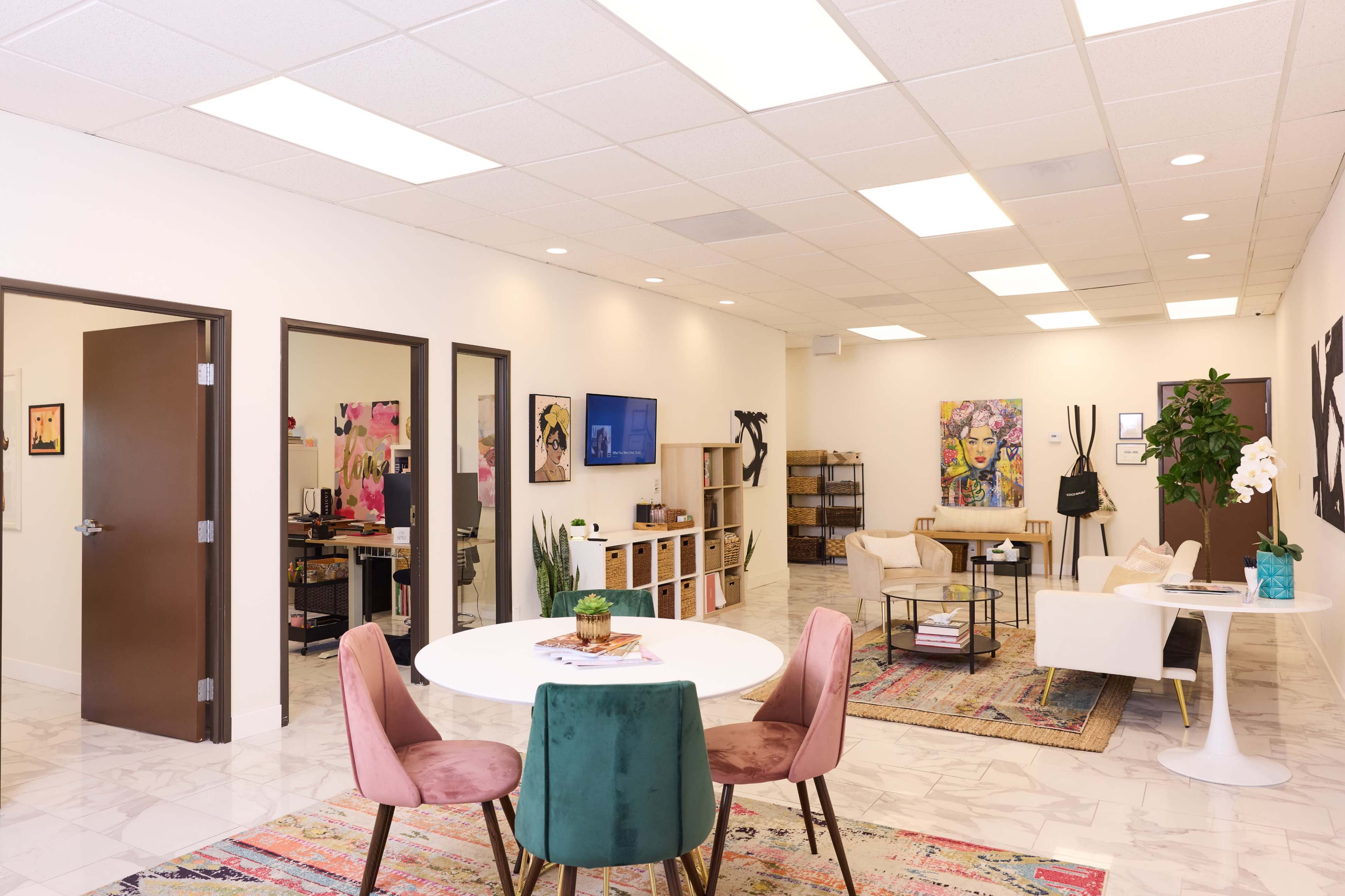 A modern office space features a round table with pink chairs, a lounge area with a white sofa, and multiple art pieces on the walls, illuminated by bright overhead lighting.