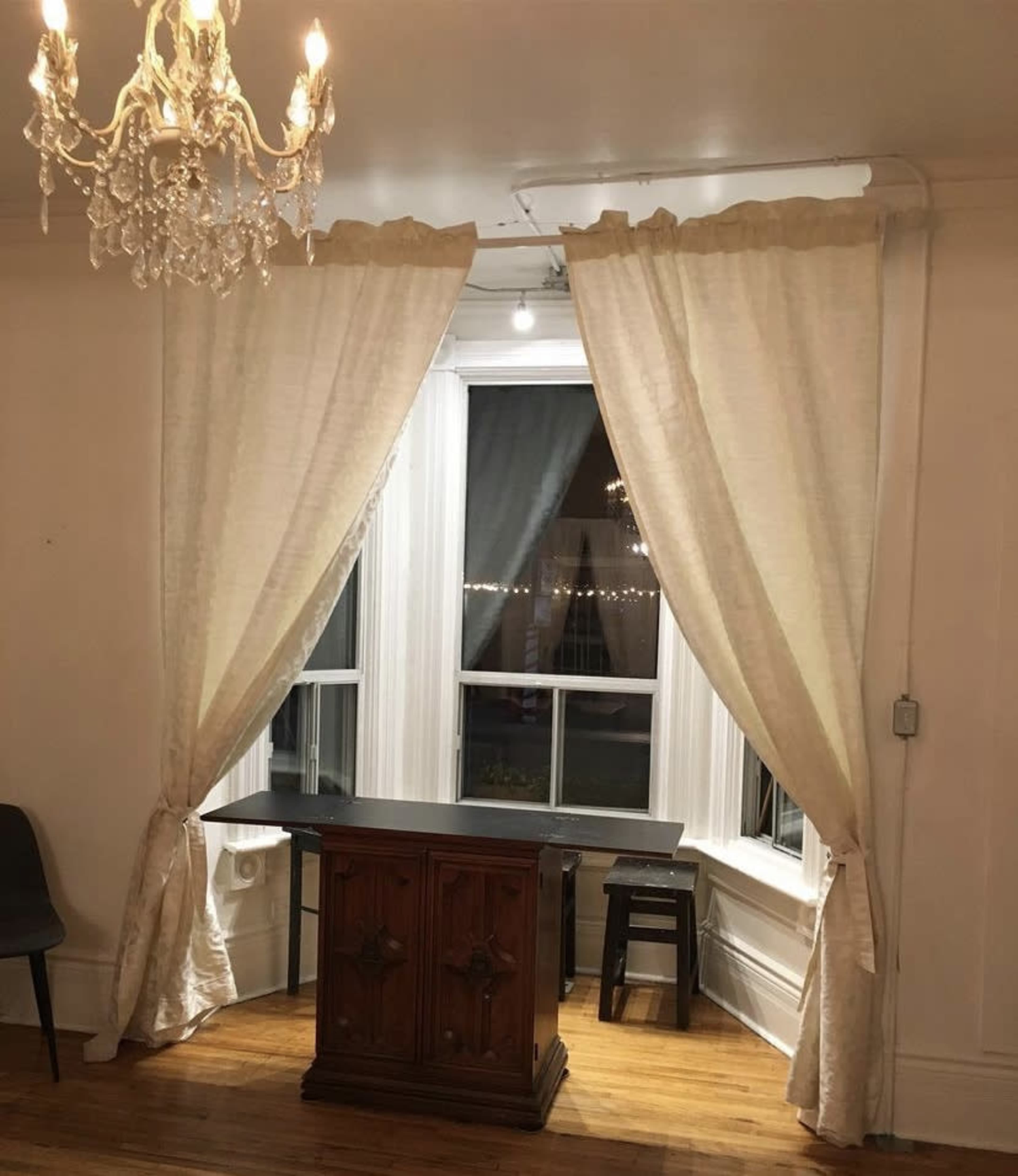 A room with a chandelier, white curtains framing a large window, and a small wooden table with a stool in front of it.
