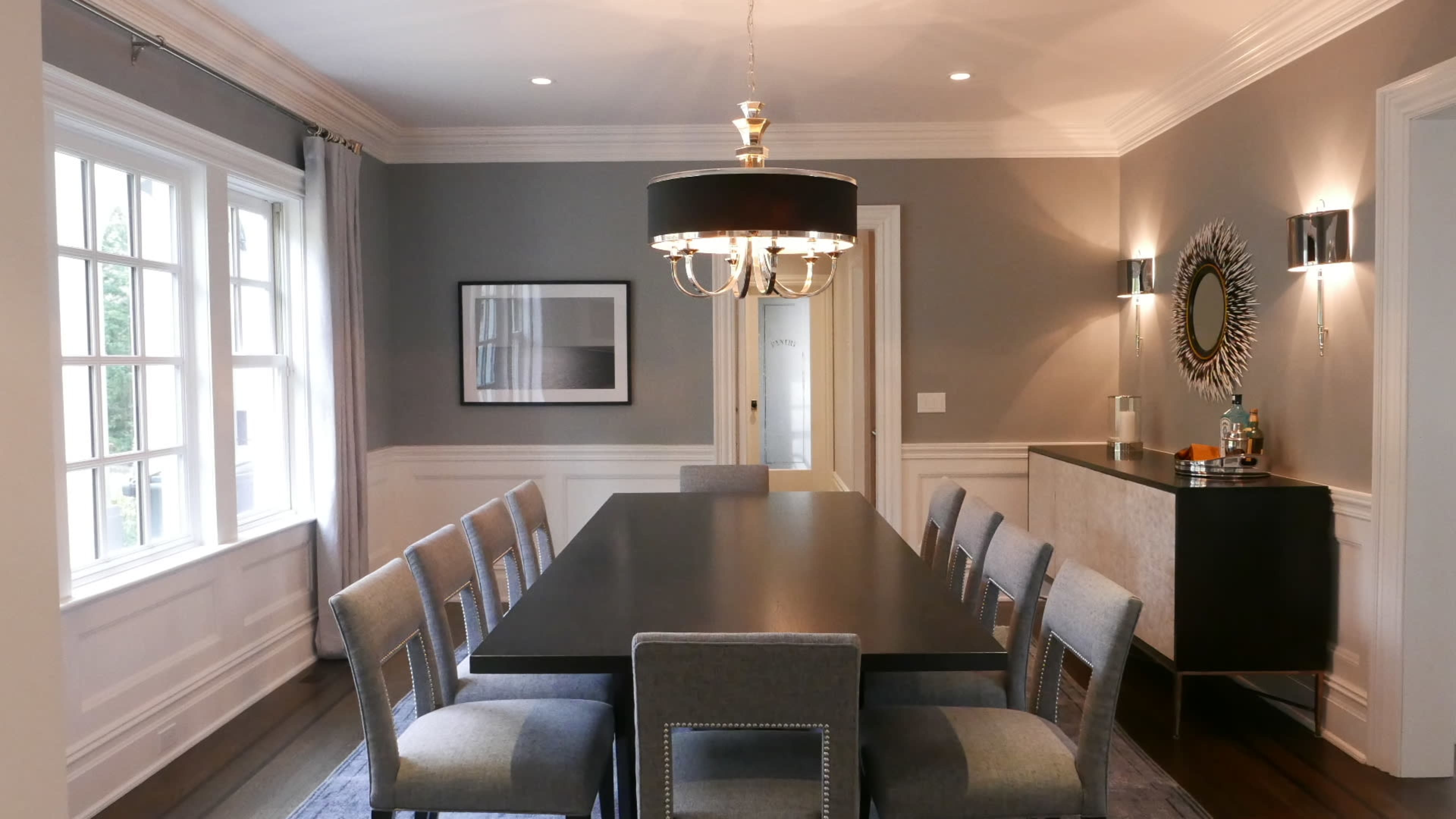 The image shows a dining room with a long table surrounded by eight upholstered chairs, a chandelier overhead, and windows providing natural light.