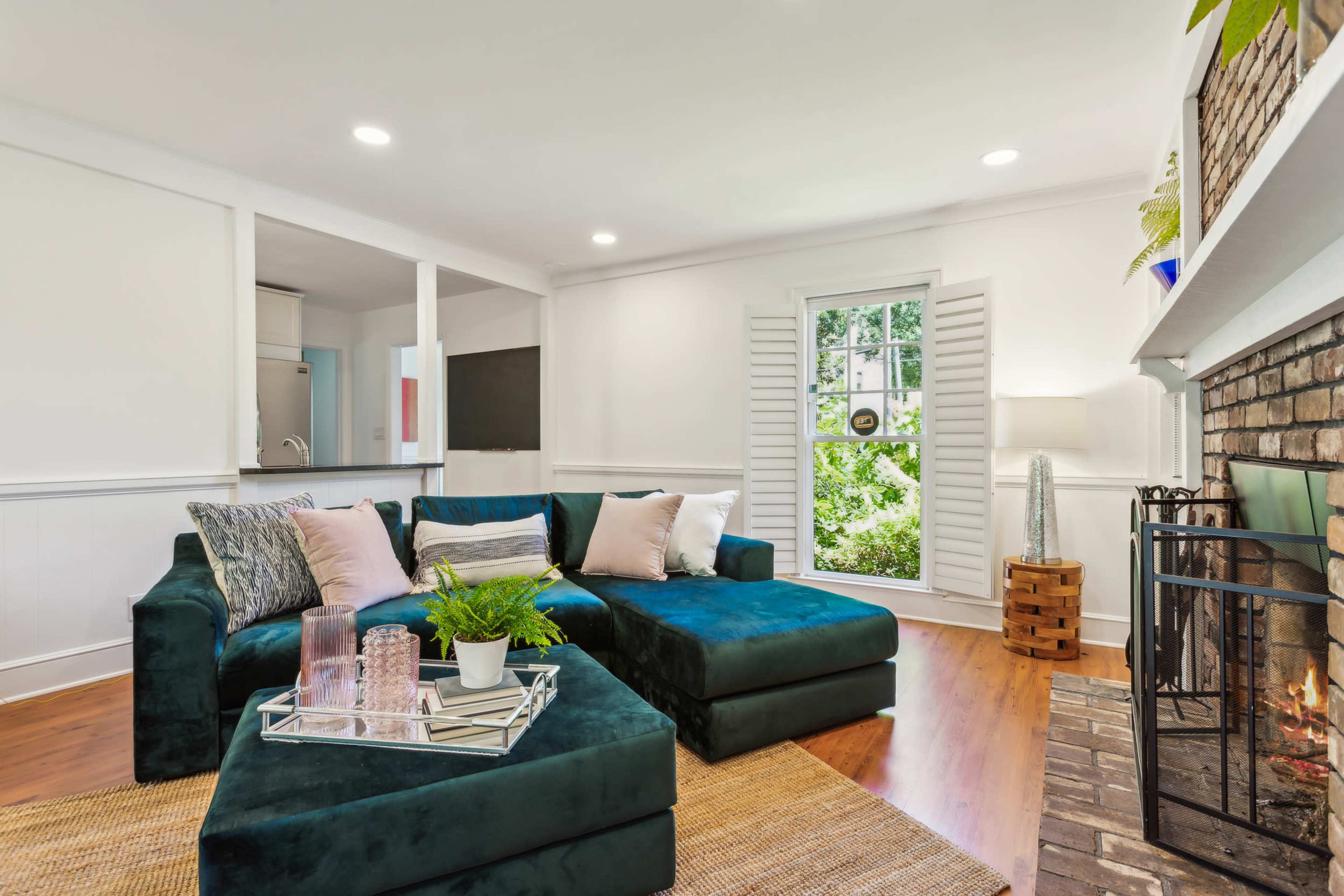 The image shows a cozy living room featuring a green sectional sofa, a brick fireplace, and a window with white shutters, surrounded by natural light.