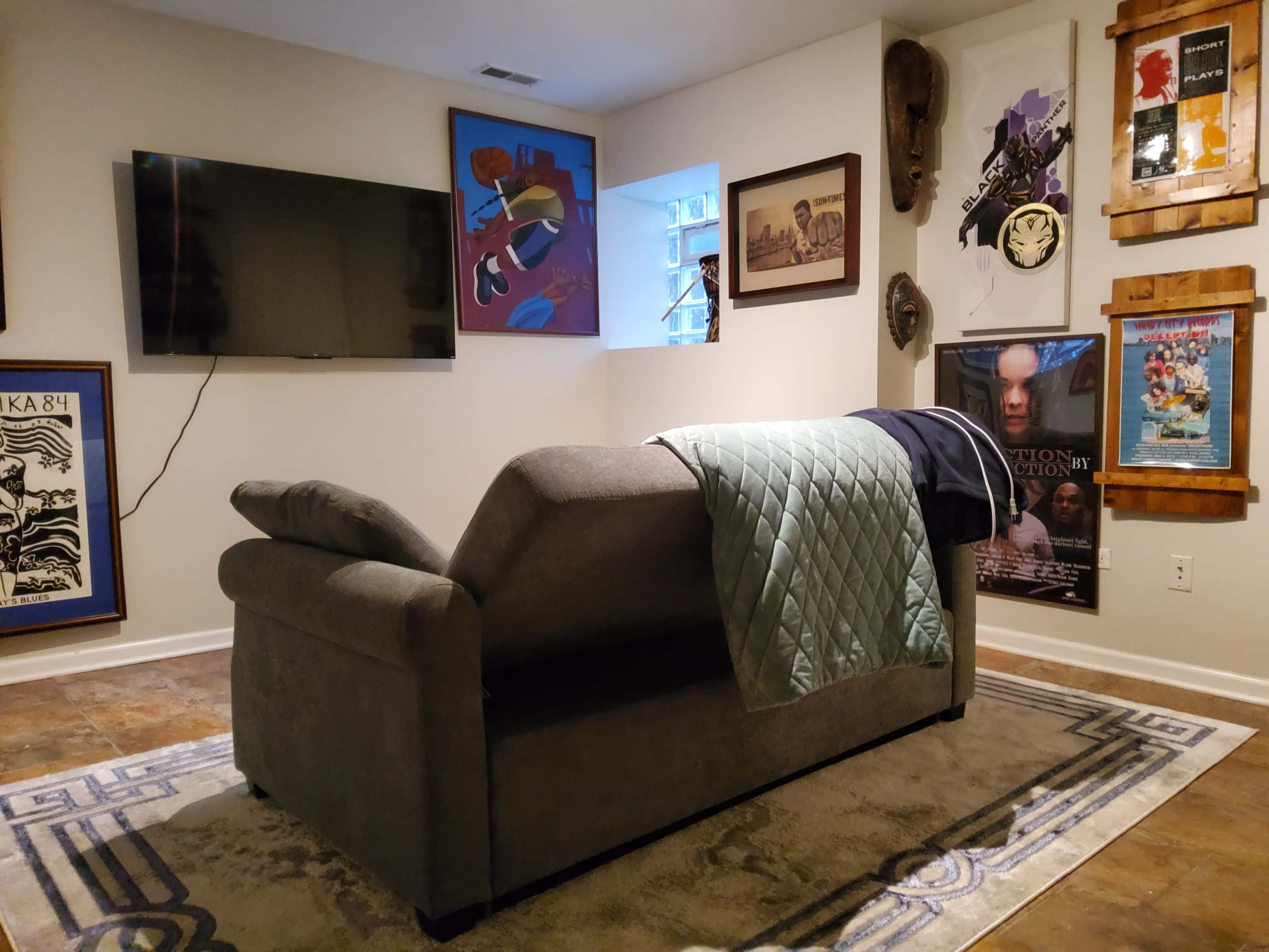 The image shows a cozy living room with a gray sectional couch, a television mounted on the wall, and various framed artworks and masks displayed on the adjacent walls.