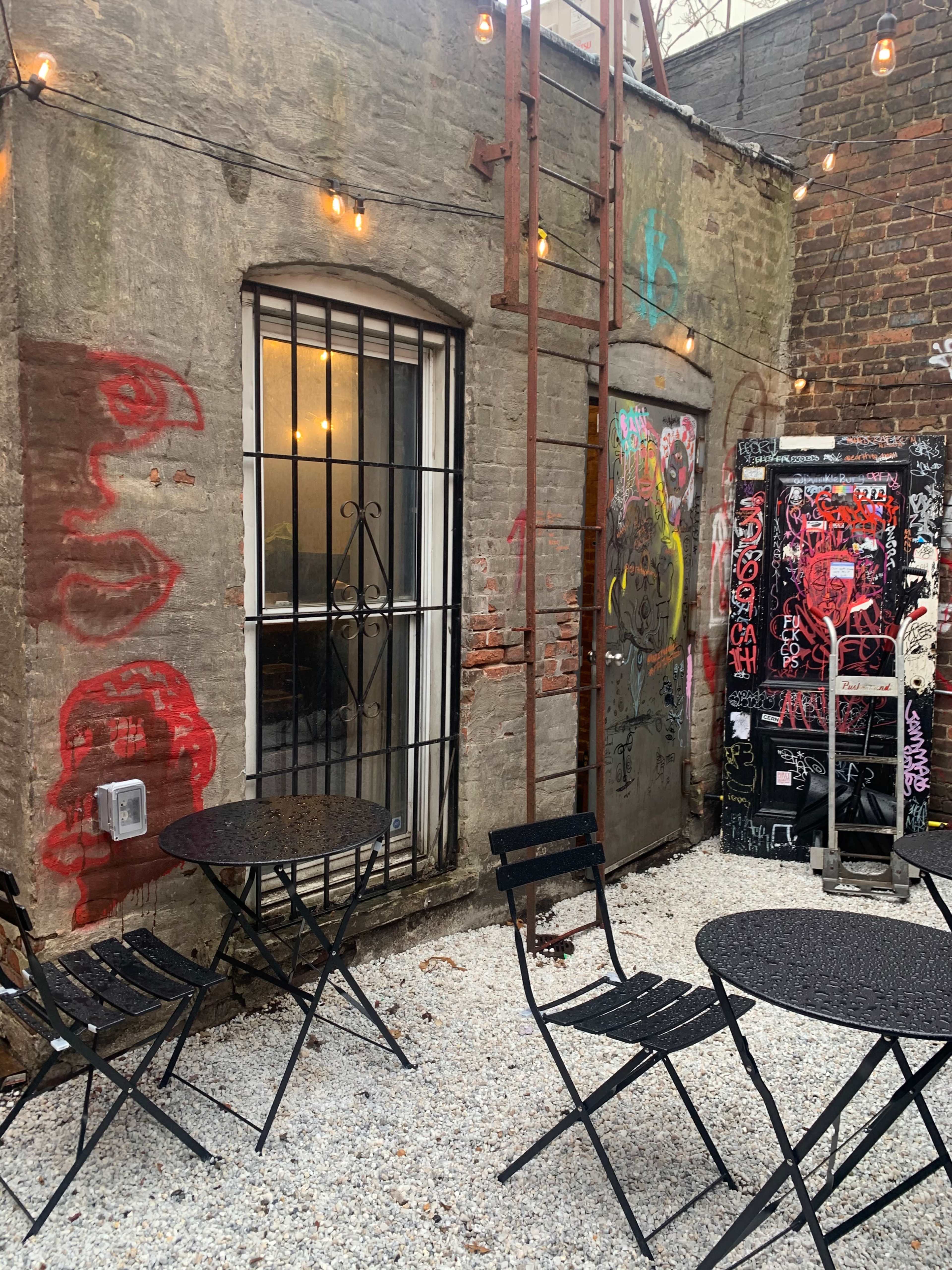 A small outdoor seating area with black metal tables and chairs is set against a graffiti-covered wall and an adjacent building with a barred window.