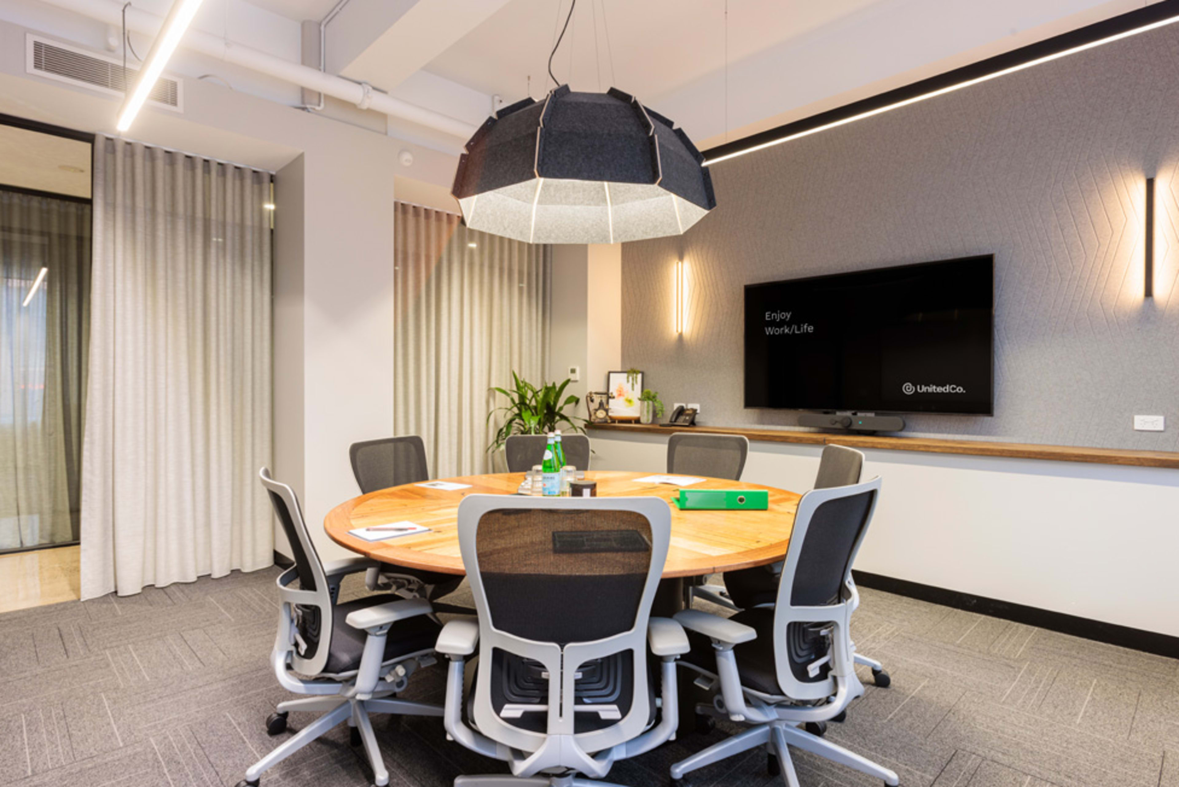 A modern conference room with a circular wooden table surrounded by ergonomic chairs, a large pendant light overhead, and a TV mounted on the wall.