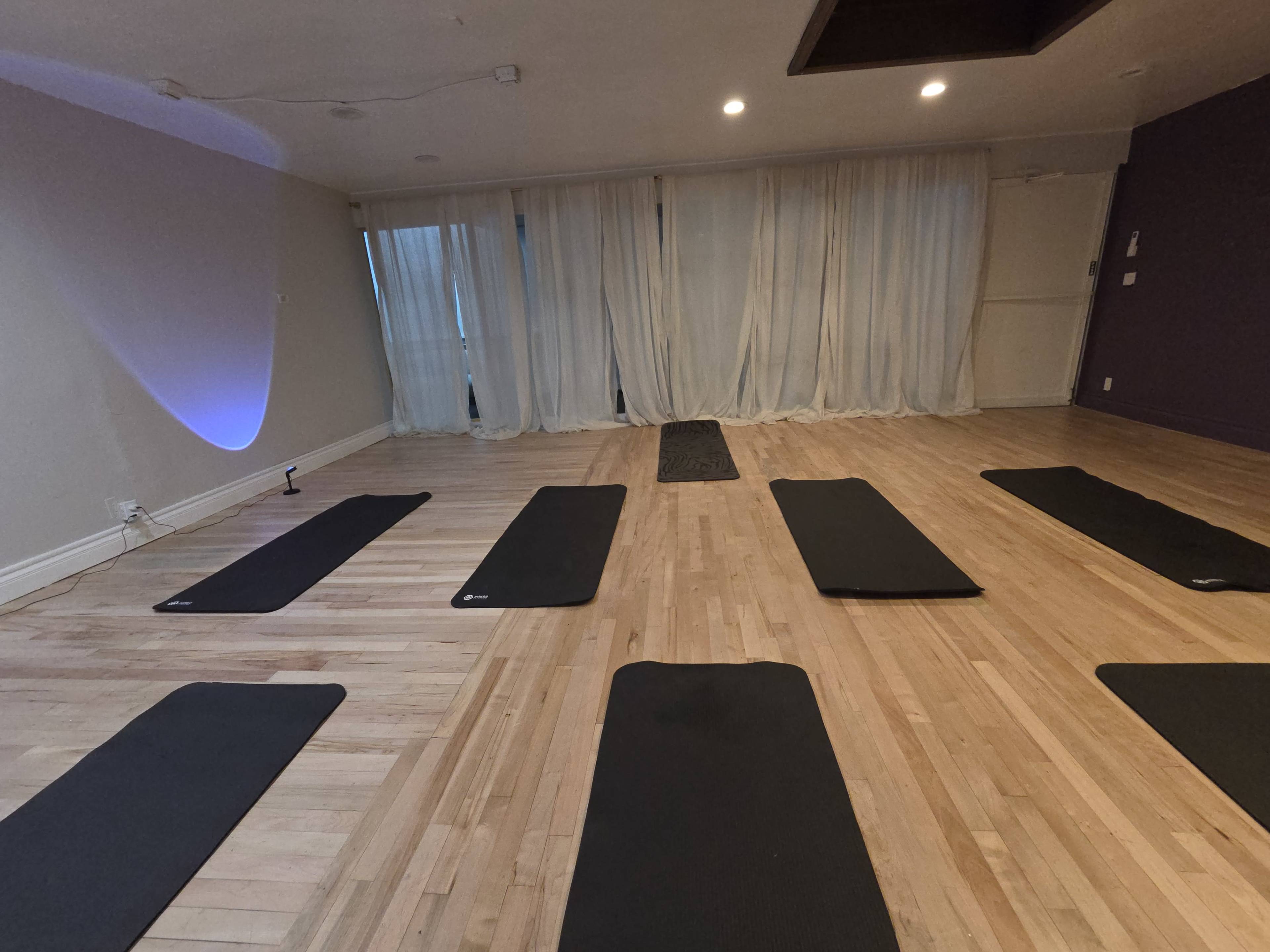 The image shows a yoga studio with wooden floors and several black yoga mats arranged on the ground, facing a wall of sheer white curtains.