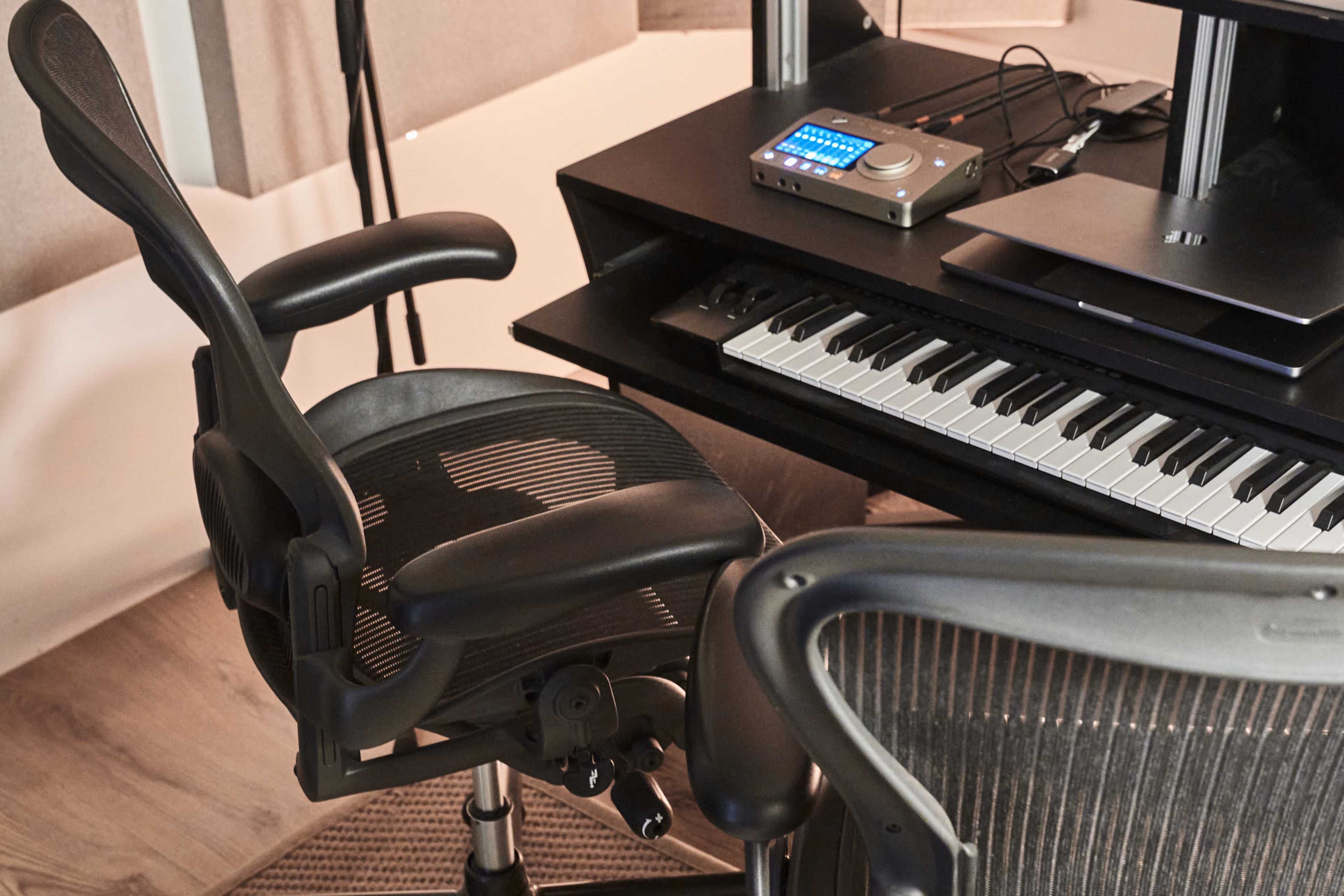 A black ergonomic chair is positioned beside a workstation featuring a keyboard and an audio interface.