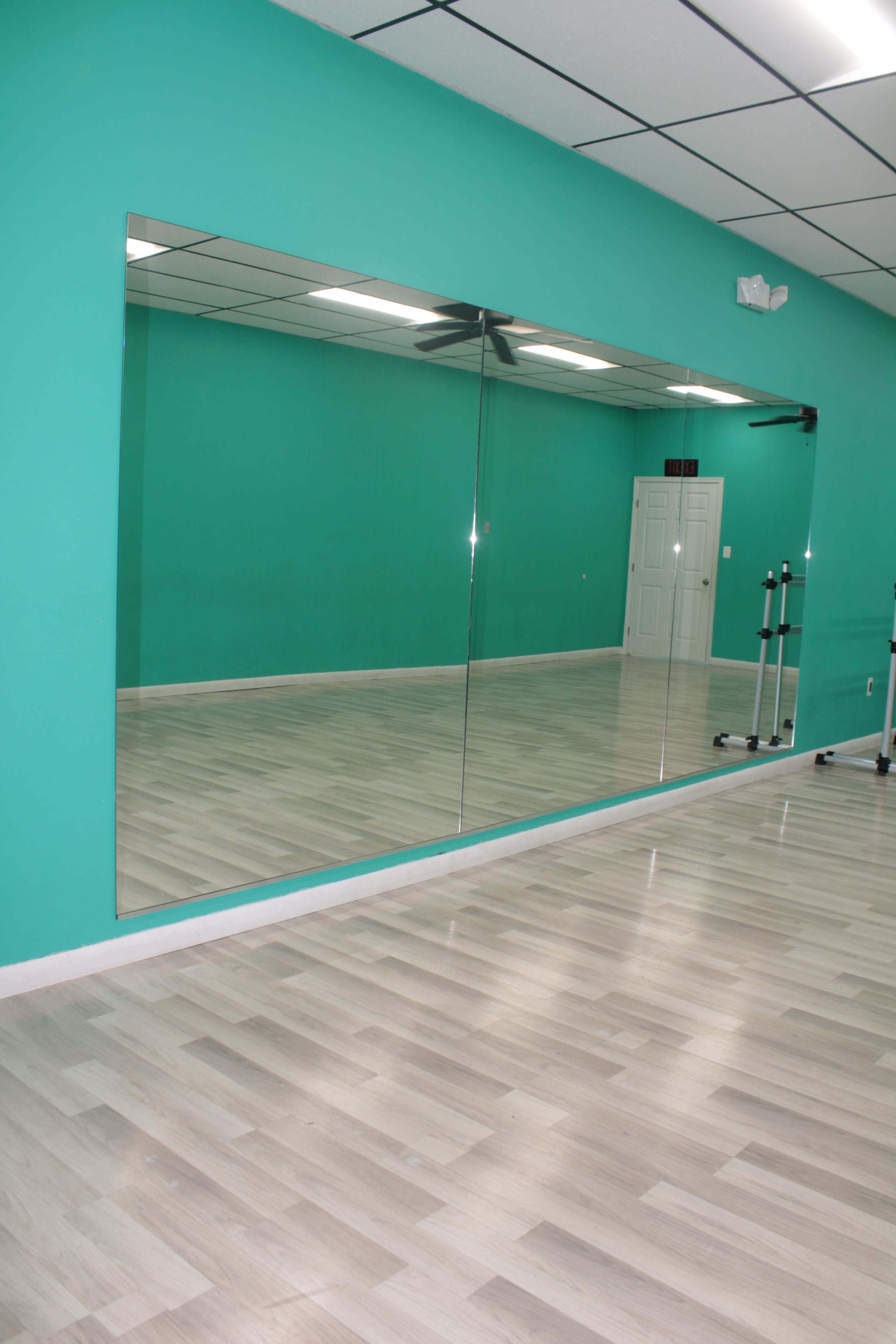 A dance studio with a large, wall-mounted mirror reflecting a turquoise wall and a light wooden floor.