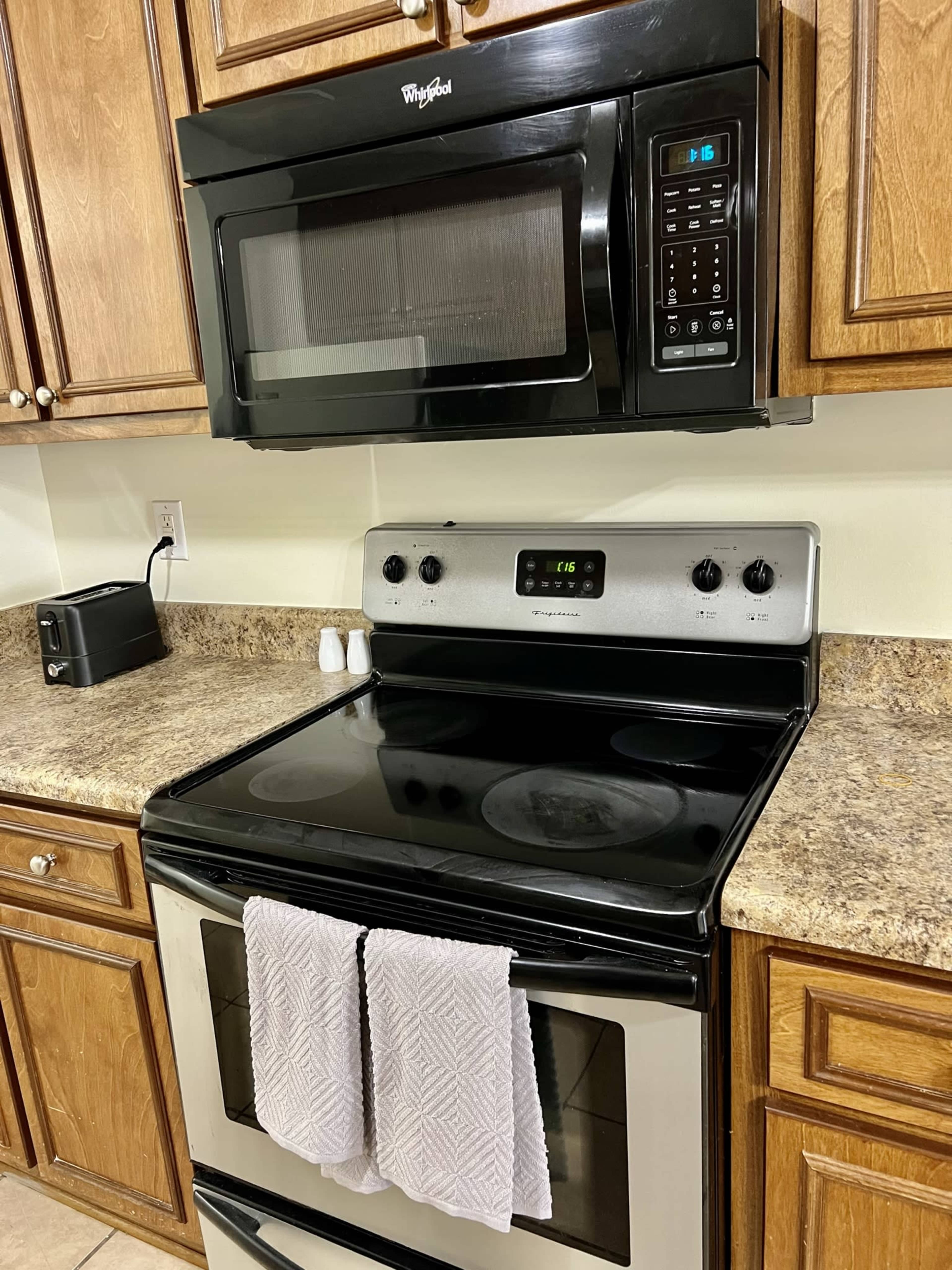 A black microwave is mounted above a stainless steel stove on a granite countertop, surrounded by wooden cabinetry.
