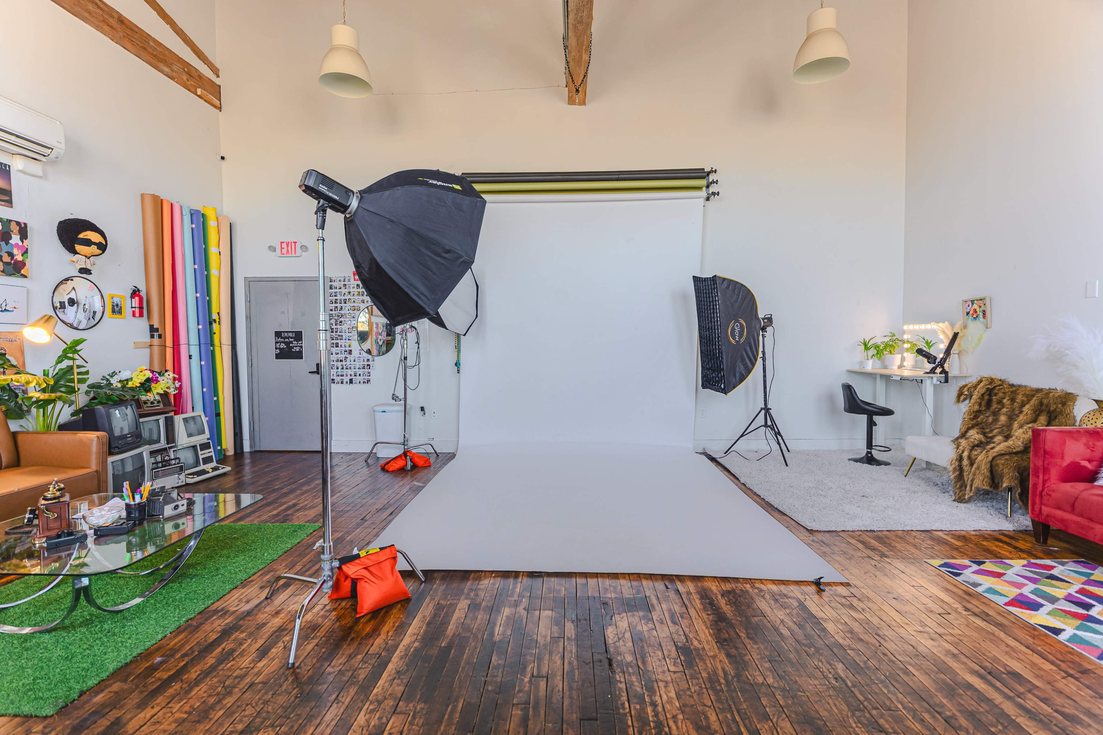 The image shows a spacious photography studio equipped with a gray backdrop, lighting equipment, and various props, alongside a seating area with colorful décor.