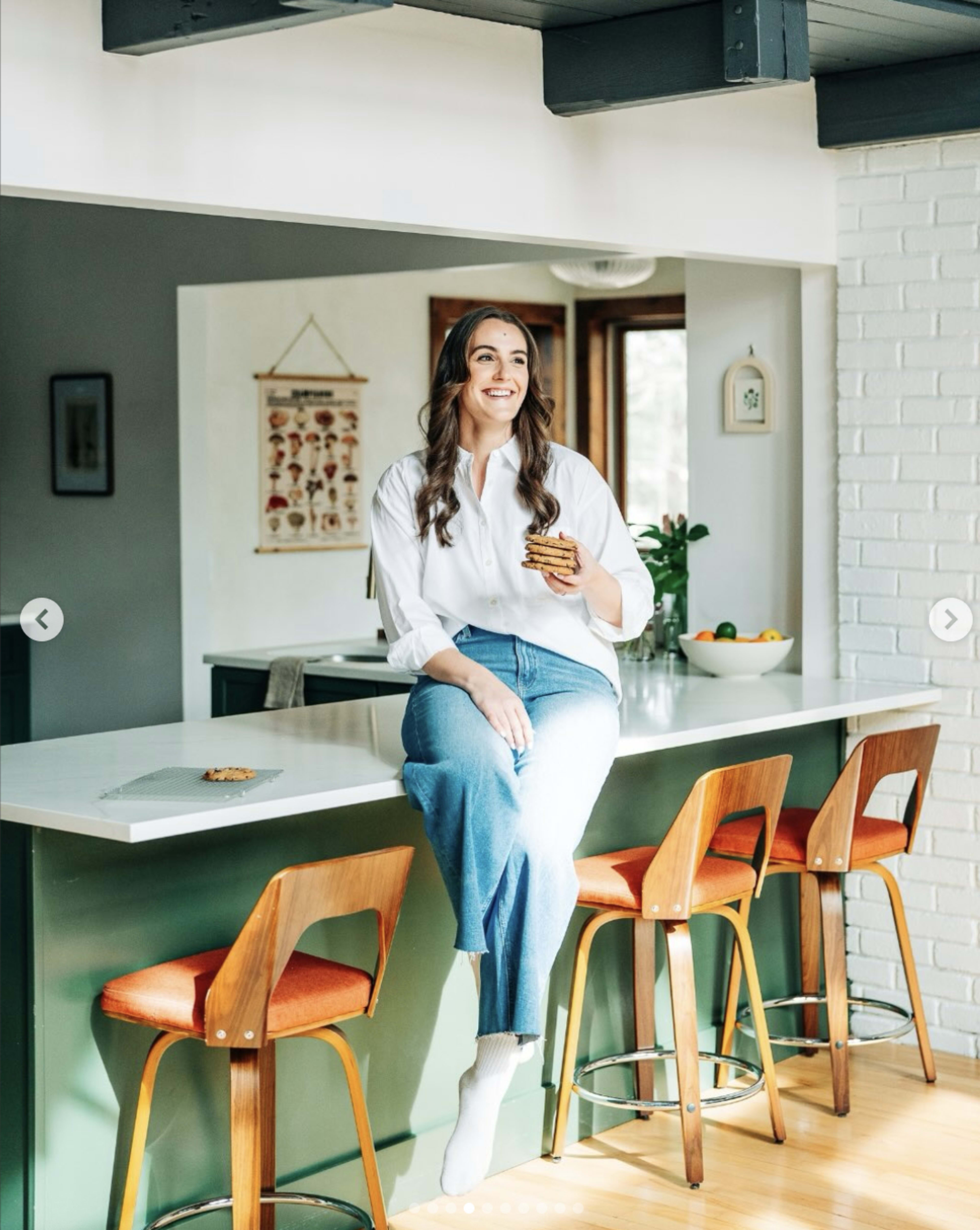 A woman sits on a kitchen counter, smiling while holding a snack, with modern bar stools and a bright, airy kitchen backdrop.
