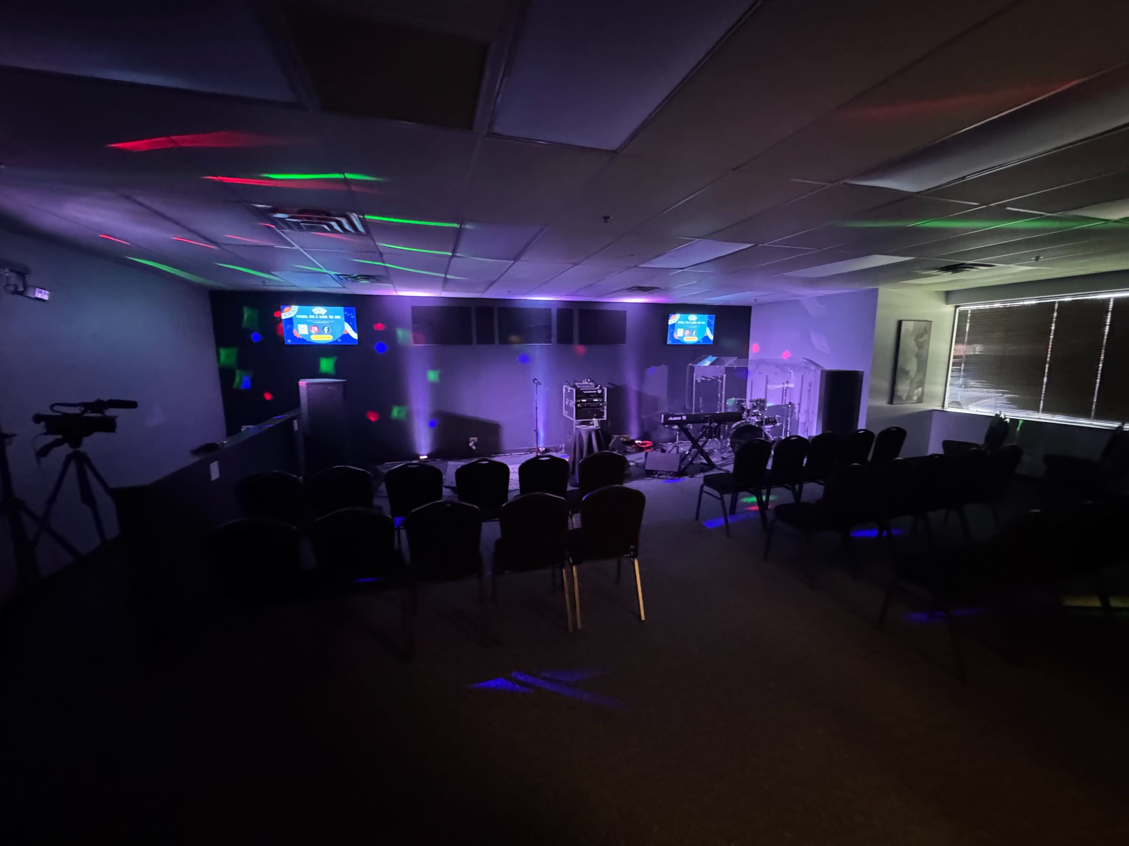The image shows a dimly lit room set up for a performance, featuring a stage area with musical equipment and a row of chairs arranged for an audience.