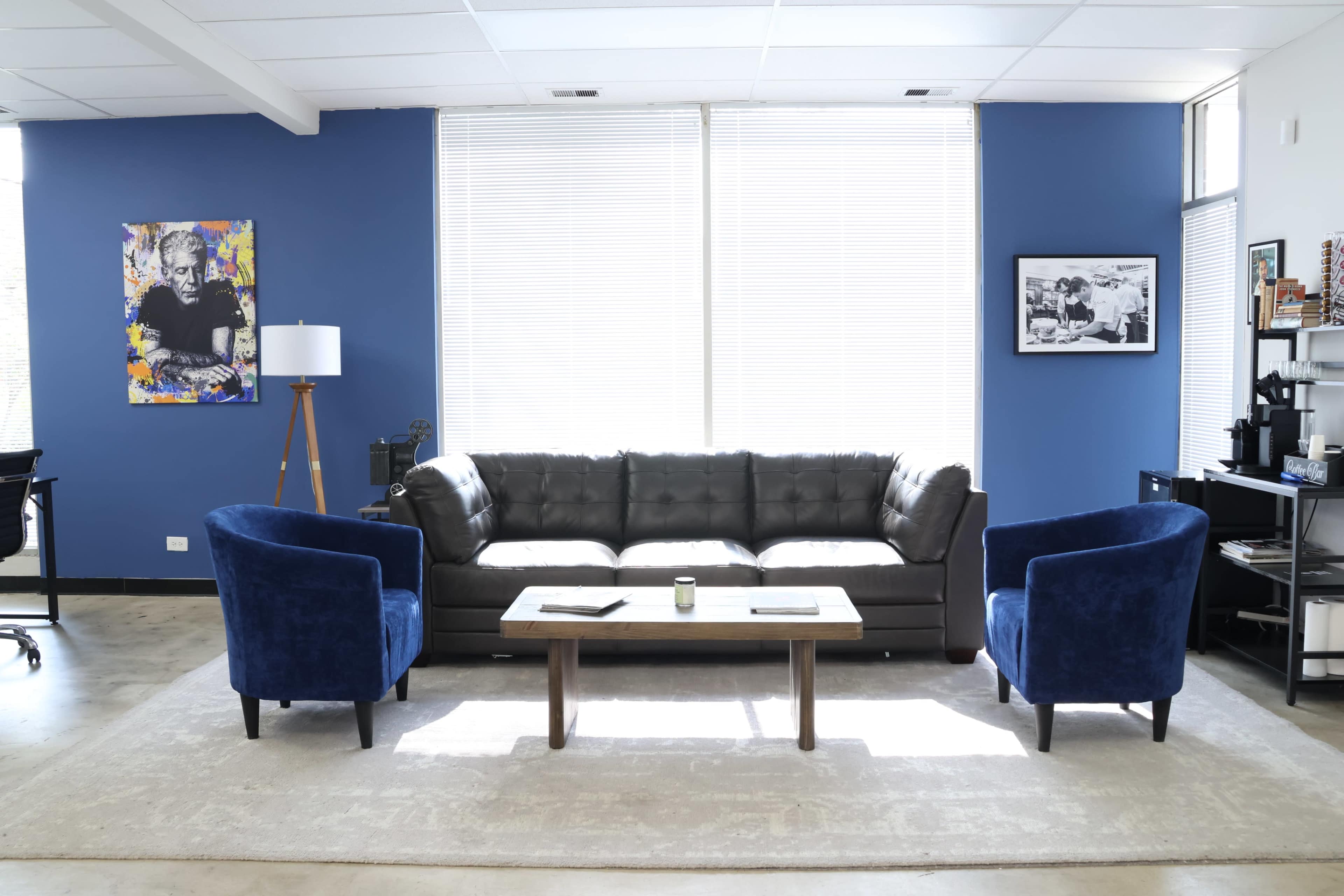 A modern lounge area with a brown leather sofa, two blue armchairs, a wooden coffee table, and large windows with white blinds.