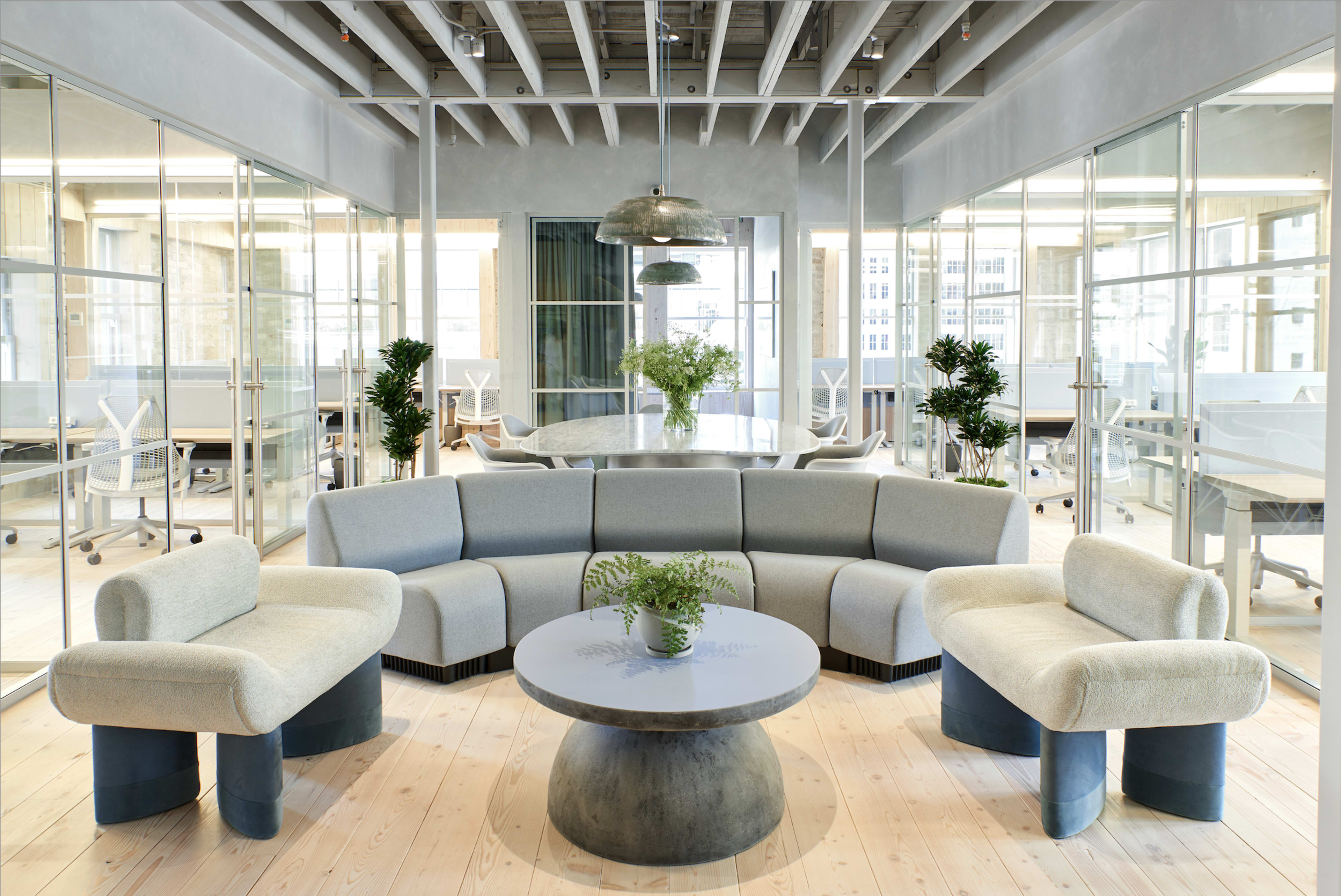 The image shows a modern office space featuring a circular gray sofa and a round coffee table, surrounded by glass-walled meeting rooms and tables with chairs in the background.