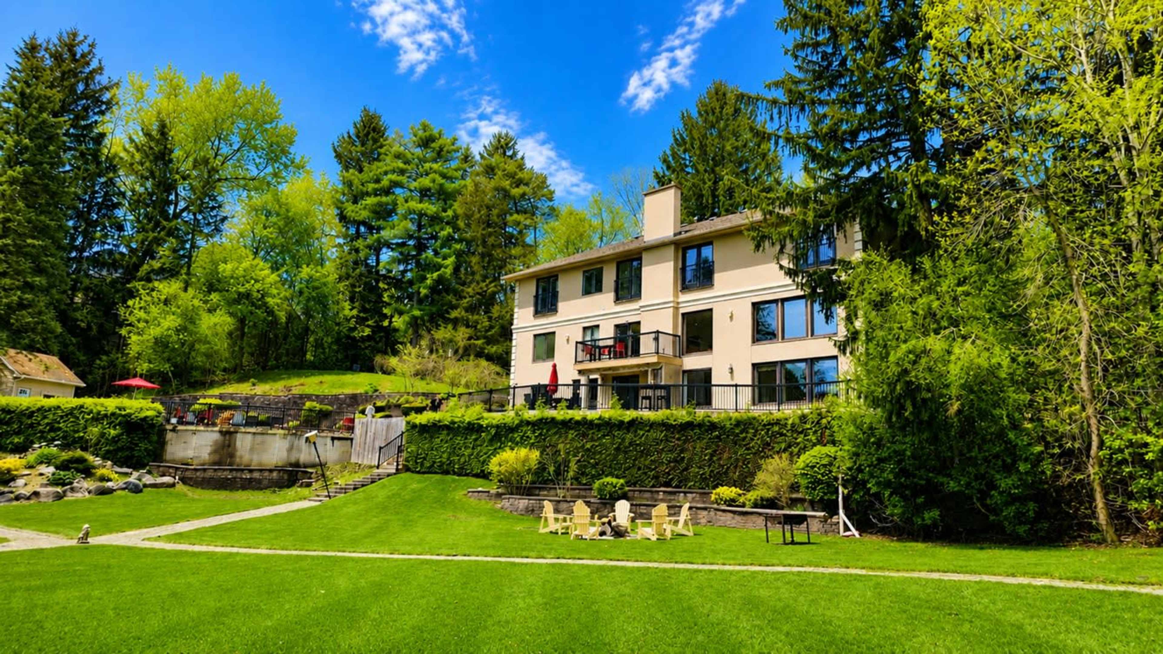 A modern house with large windows is situated on a grassy hillside surrounded by trees and a stone pathway.