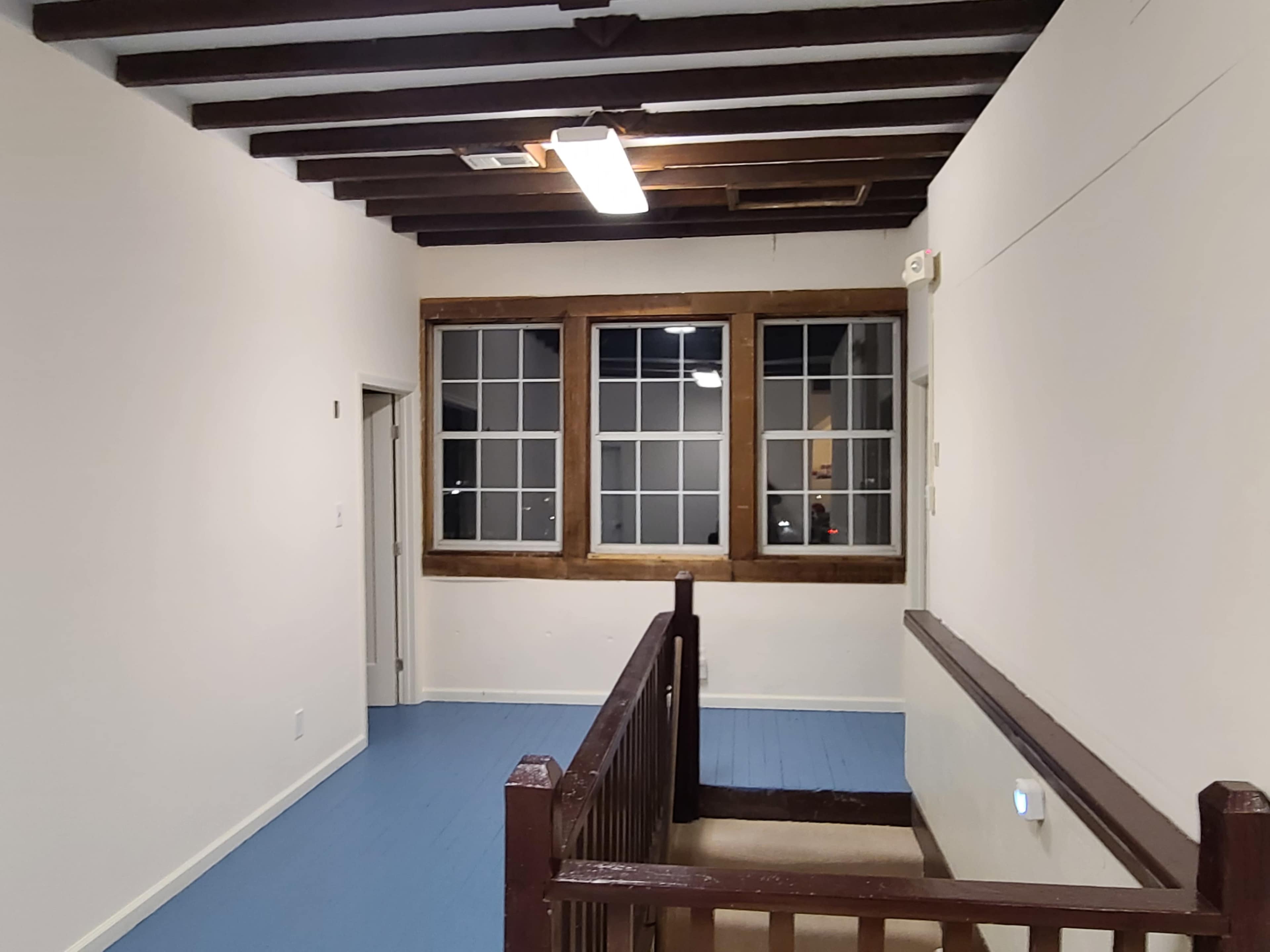A hallway with wooden beams on the ceiling, light-colored walls, and a staircase leading down to a lower level, featuring a large window with multiple panes.