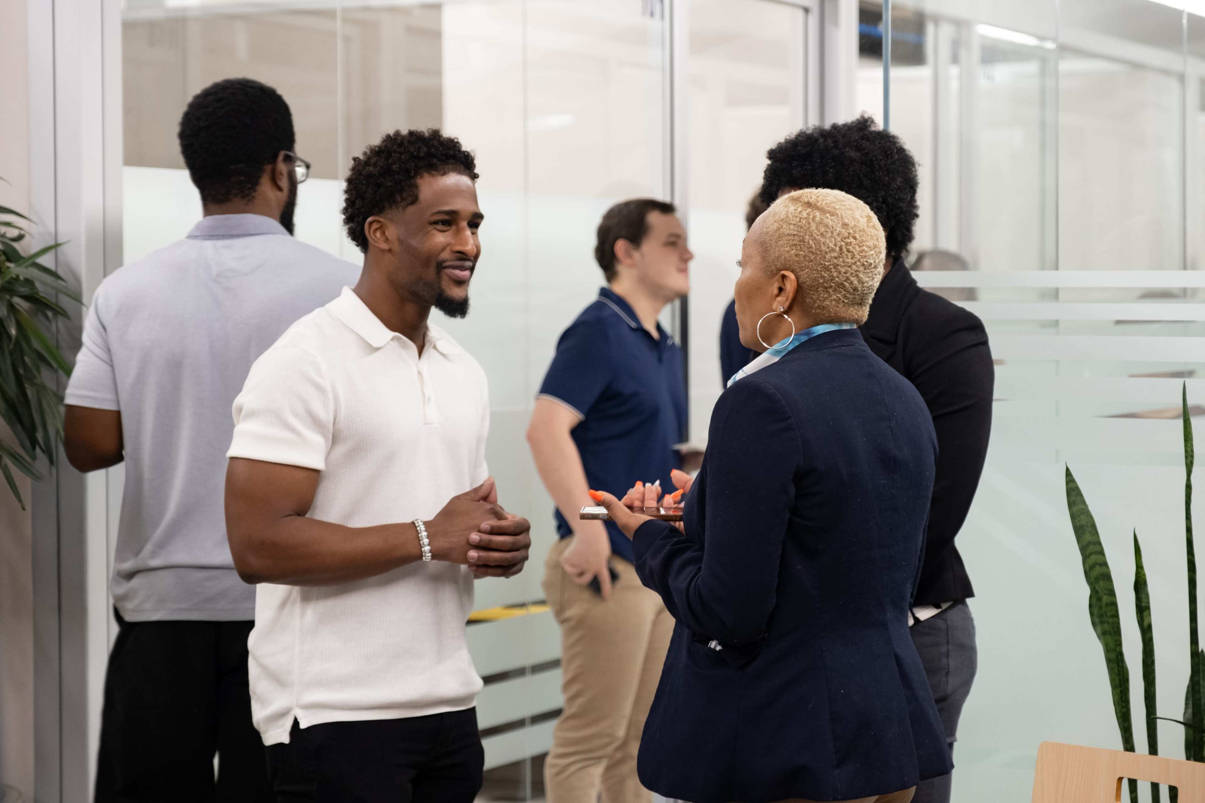 A group of professionals engages in conversation in a modern office setting, with one man speaking to a woman while others are gathered in the background.