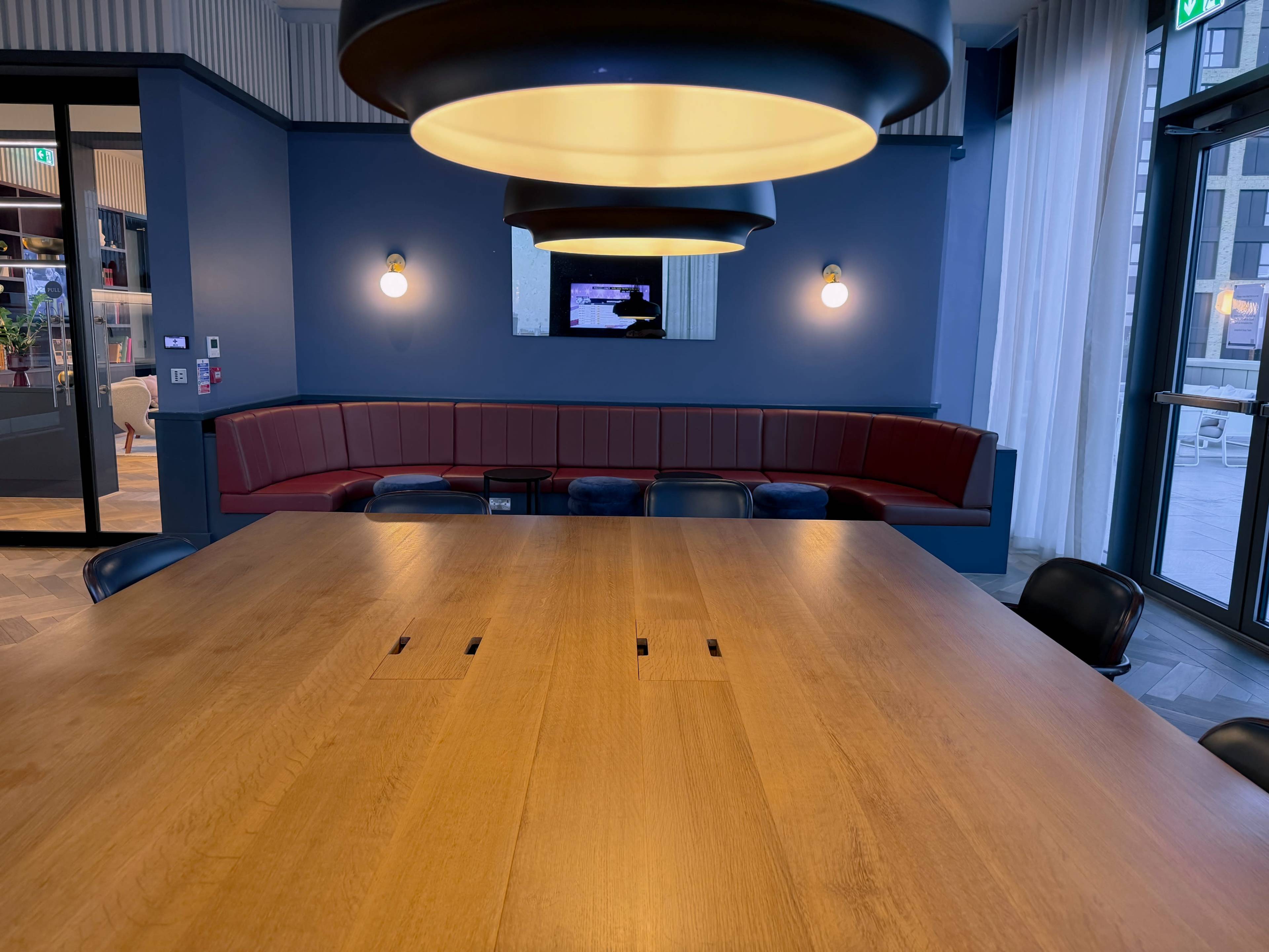 A large wooden conference table with electrical outlets is positioned in a room featuring a long red upholstered bench, wall lights, and a wall-mounted screen.