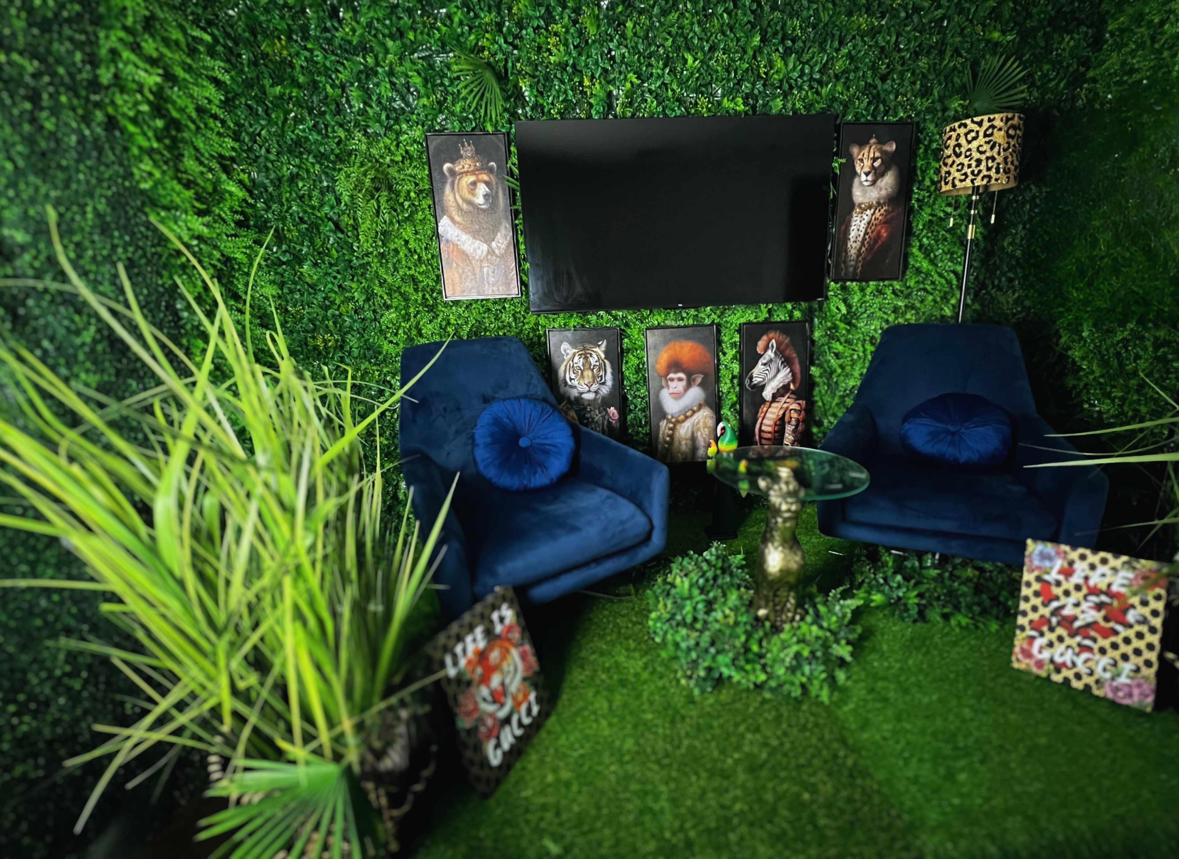 The image shows a cozy seating area with two blue chairs, a glass table, and framed animal portraits on a green, plant-covered wall.
