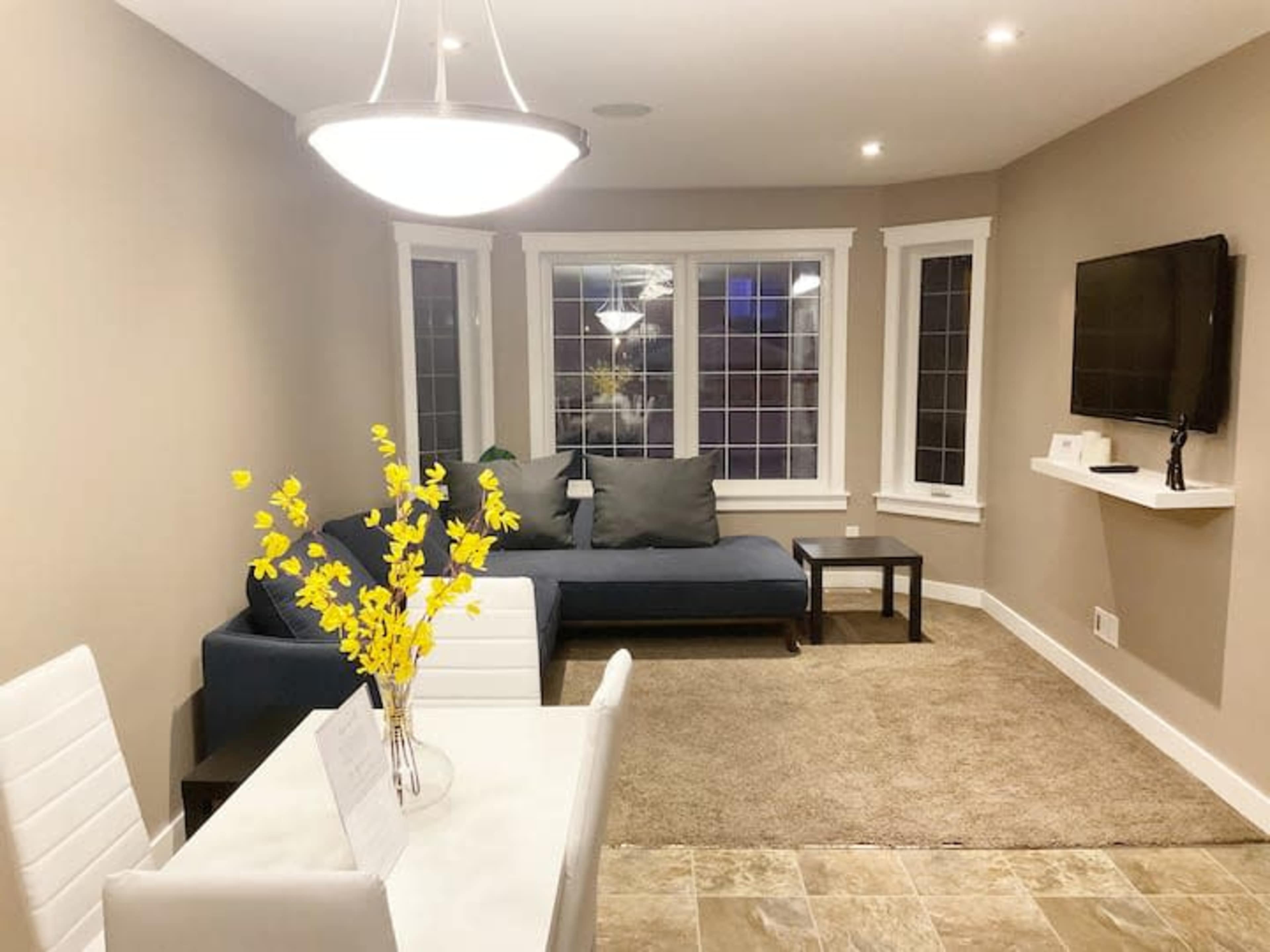 A well-lit living area featuring a blue sofa, a small table with white chairs, and a television mounted on the wall.