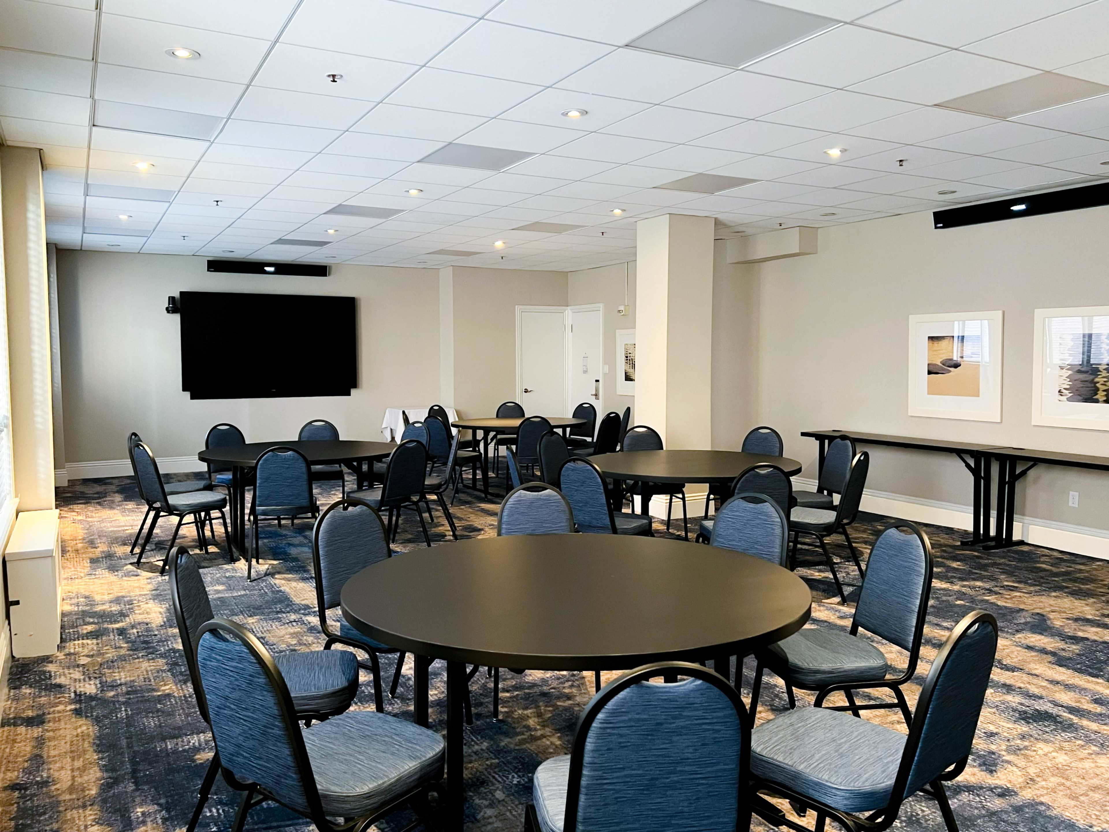 A spacious meeting room features several round tables and chairs arranged on a carpeted floor, with a large screen mounted on the wall and artwork displayed nearby.