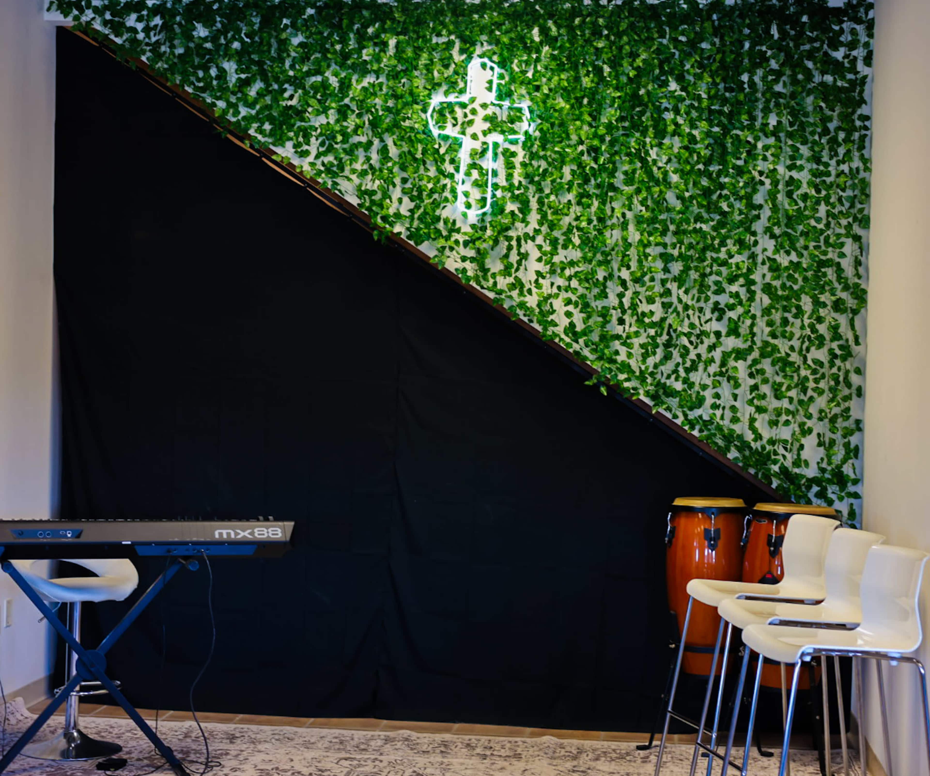 The image features a corner of a room with a black drape and a green ivy backdrop, a neon cross, a keyboard on a stand, and conga drums nearby, along with several white chairs.