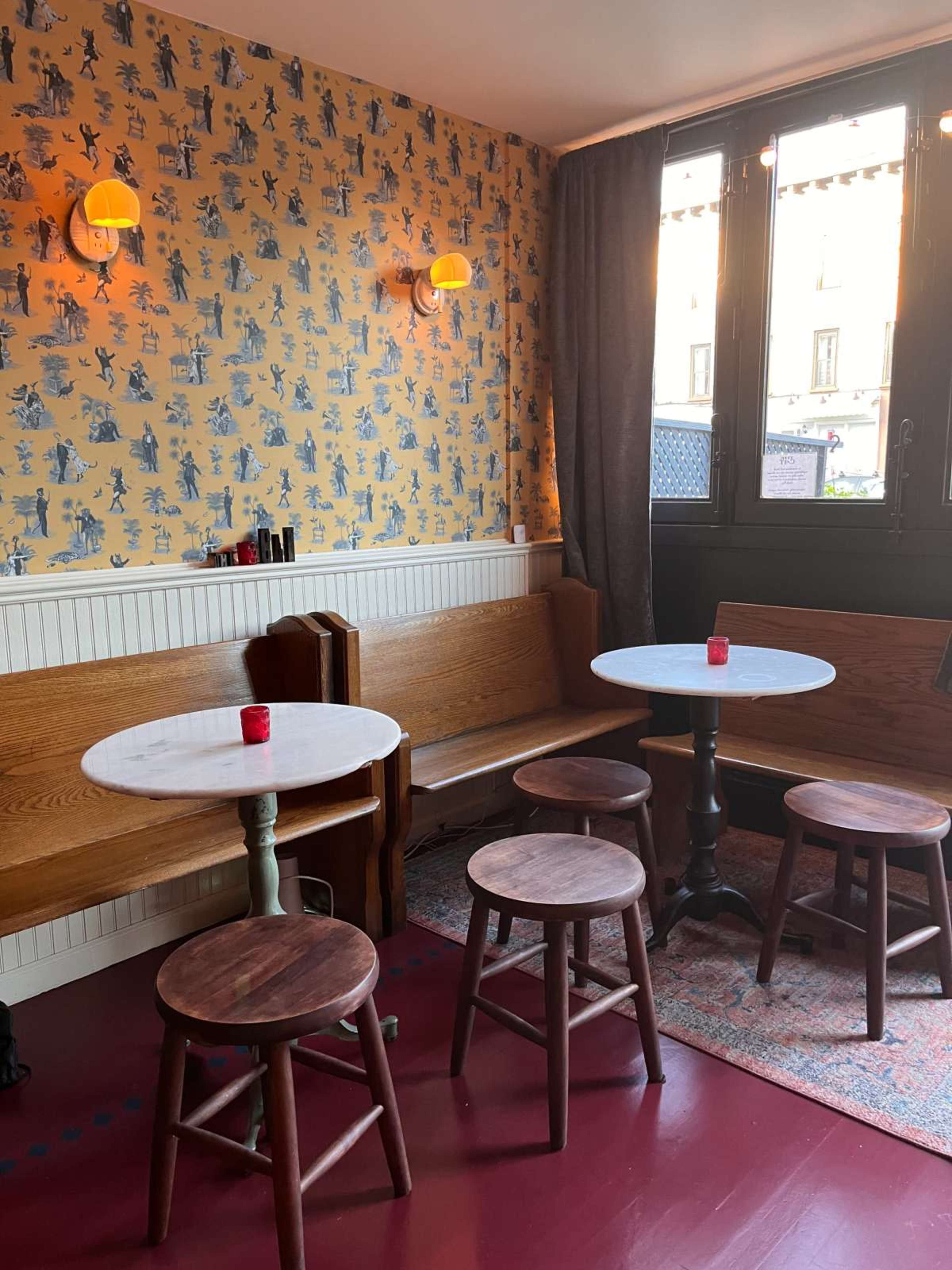 The image shows a cozy seating area with two circular marble tables surrounded by wooden stools, set against a patterned wallpaper and soft lighting.