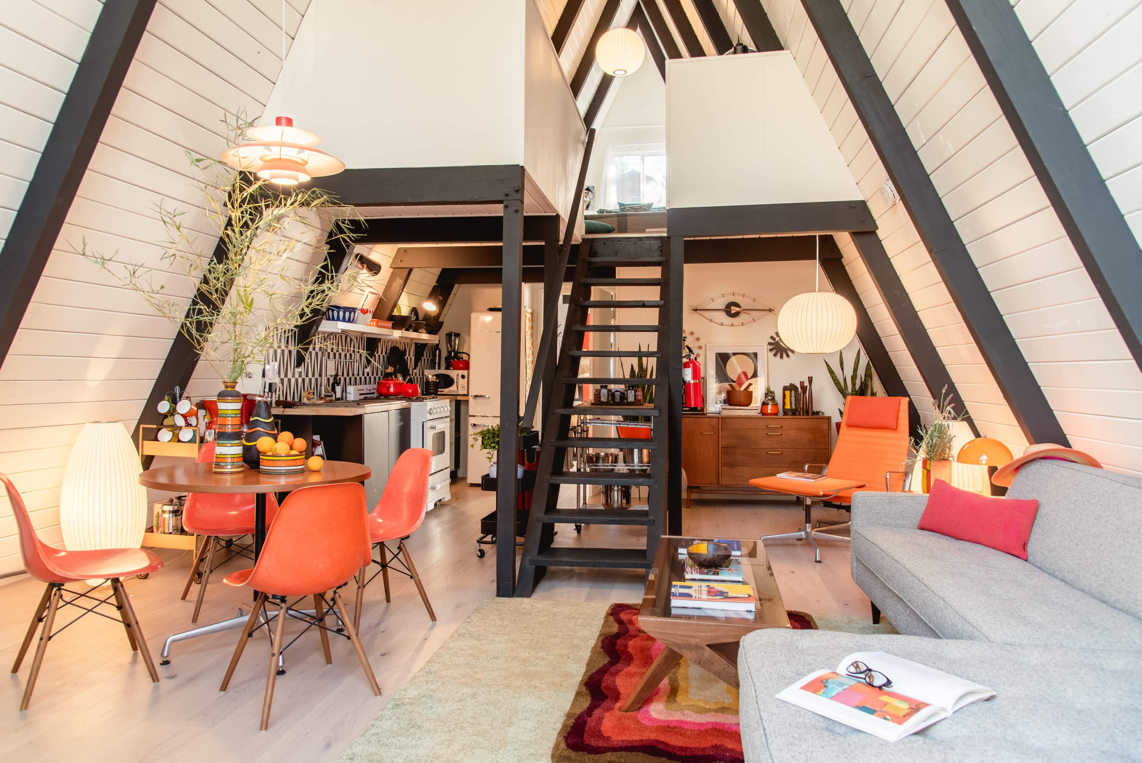 A cozy A-frame living space featuring a dining area with orange chairs, a gray sofa, and a staircase leading to a loft.