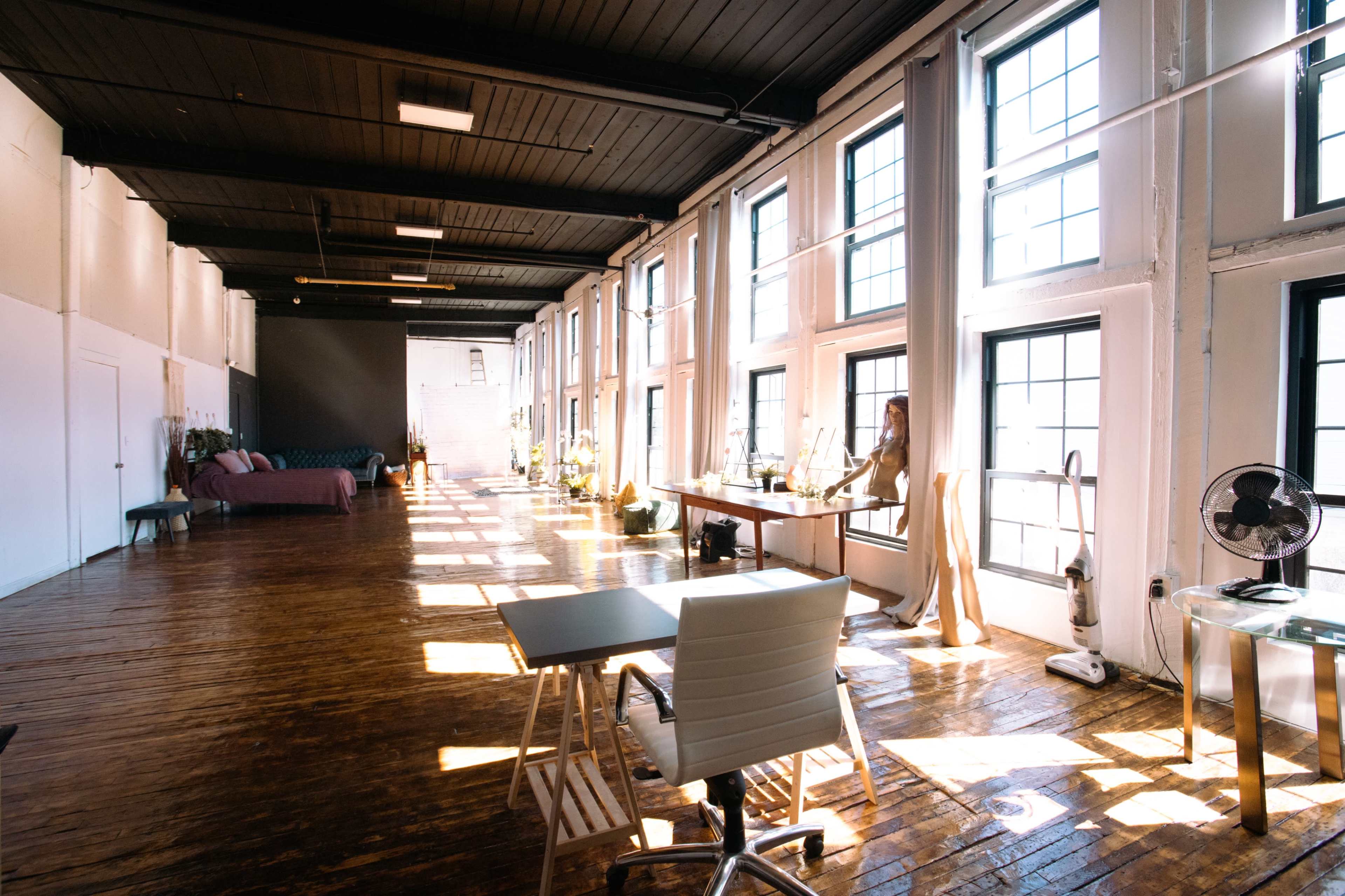 A spacious, sunlit office features large windows, wooden flooring, and minimal furnishings, including a desk and a comfortable seating area.