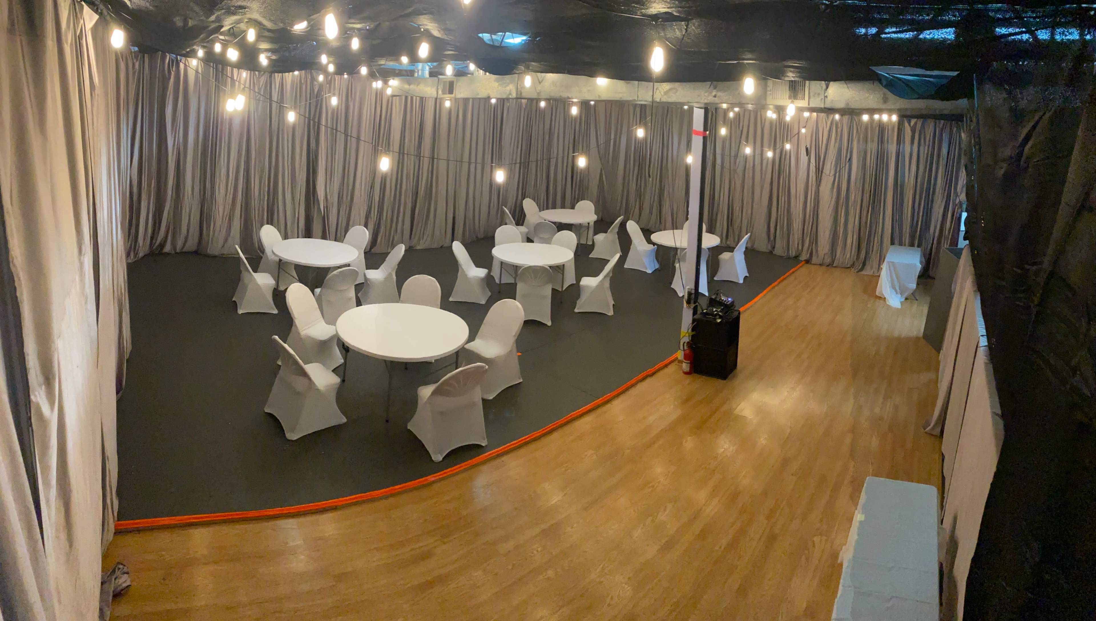 A large indoor space with several round tables covered in white cloth, surrounded by chairs, and illuminated by hanging lights.
