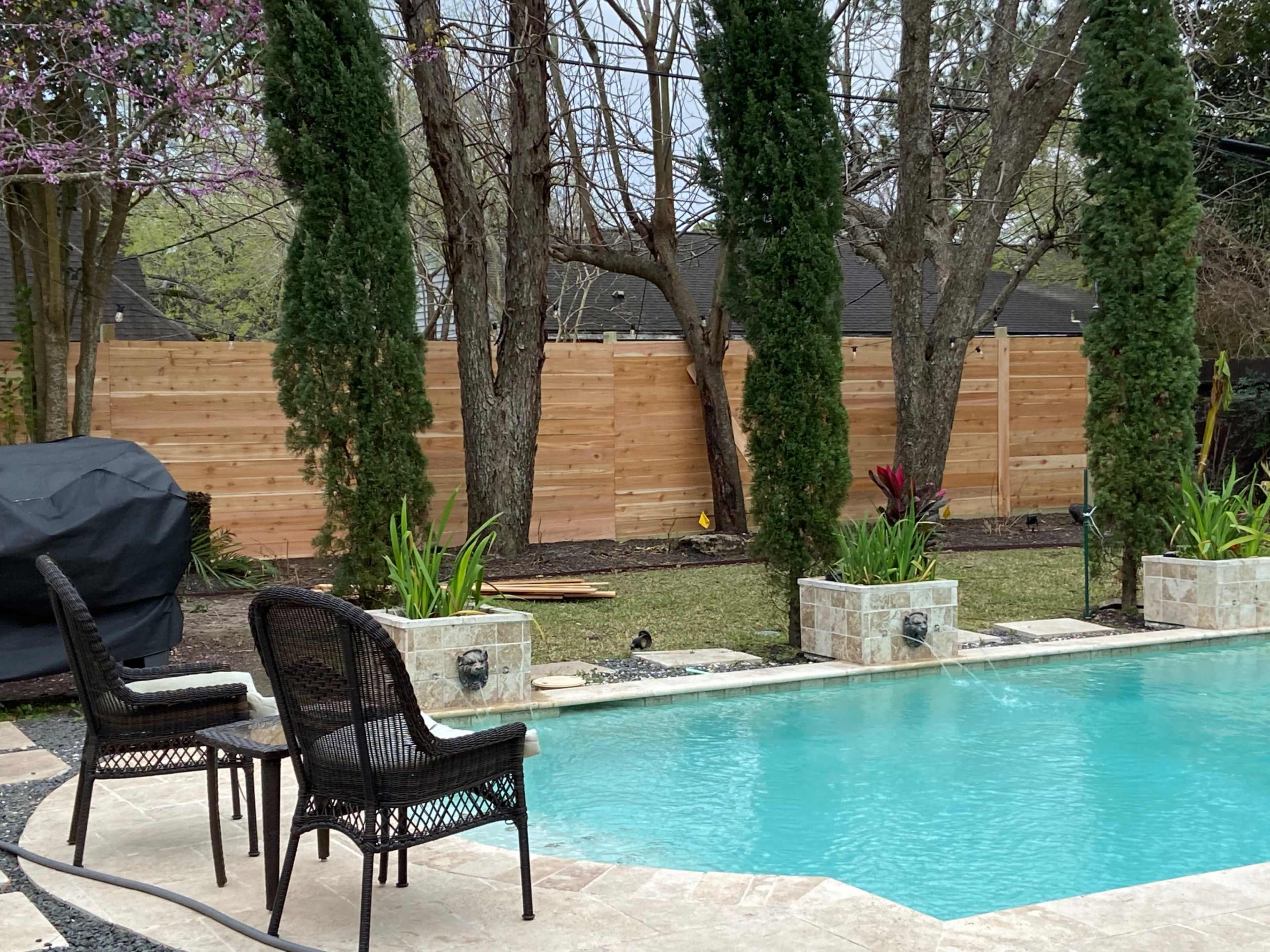 A swimming pool is surrounded by neatly arranged chairs and planters, with wooden fencing and trees in the background.