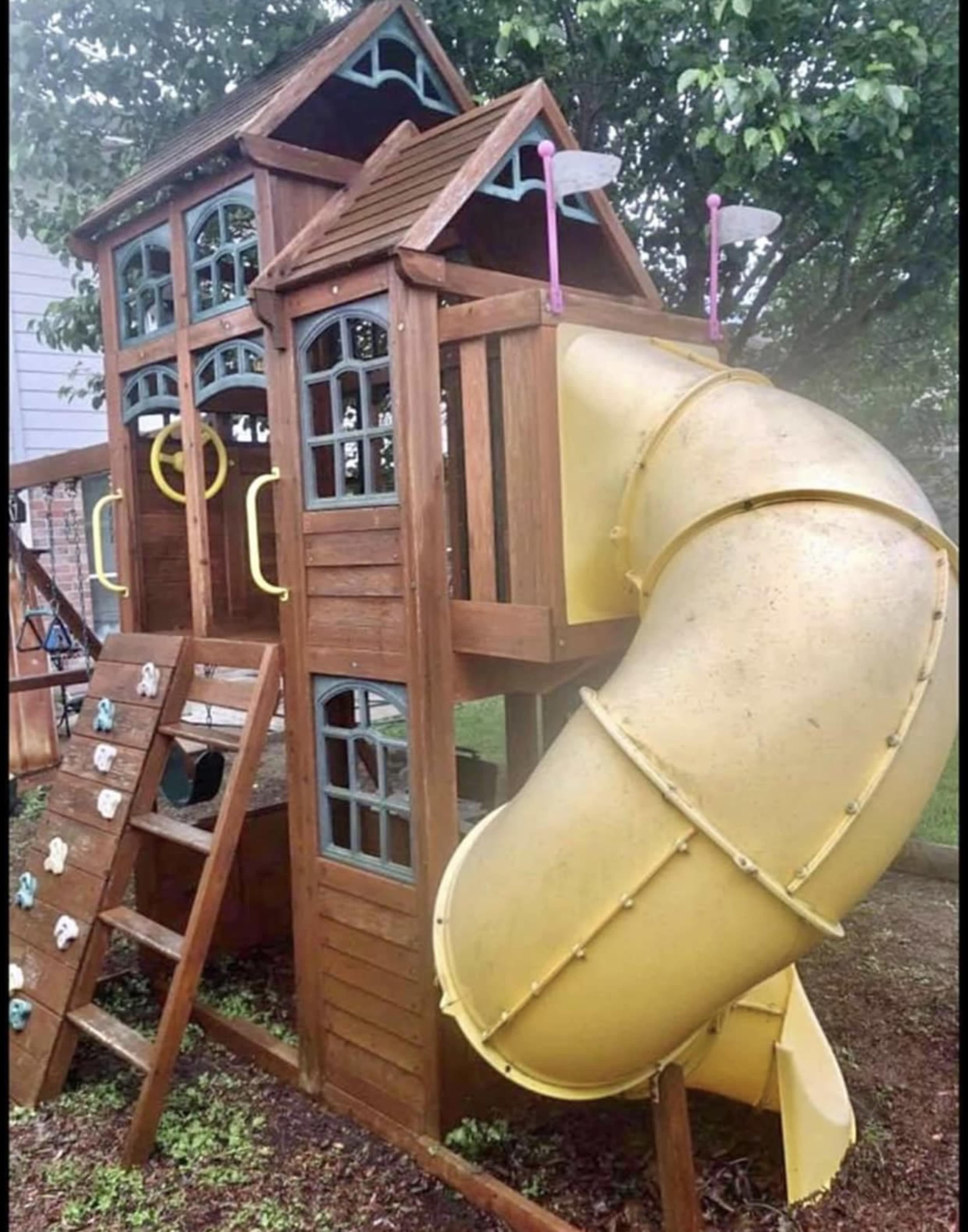 A wooden playground structure with a slide, climbing wall, and two small playhouses.