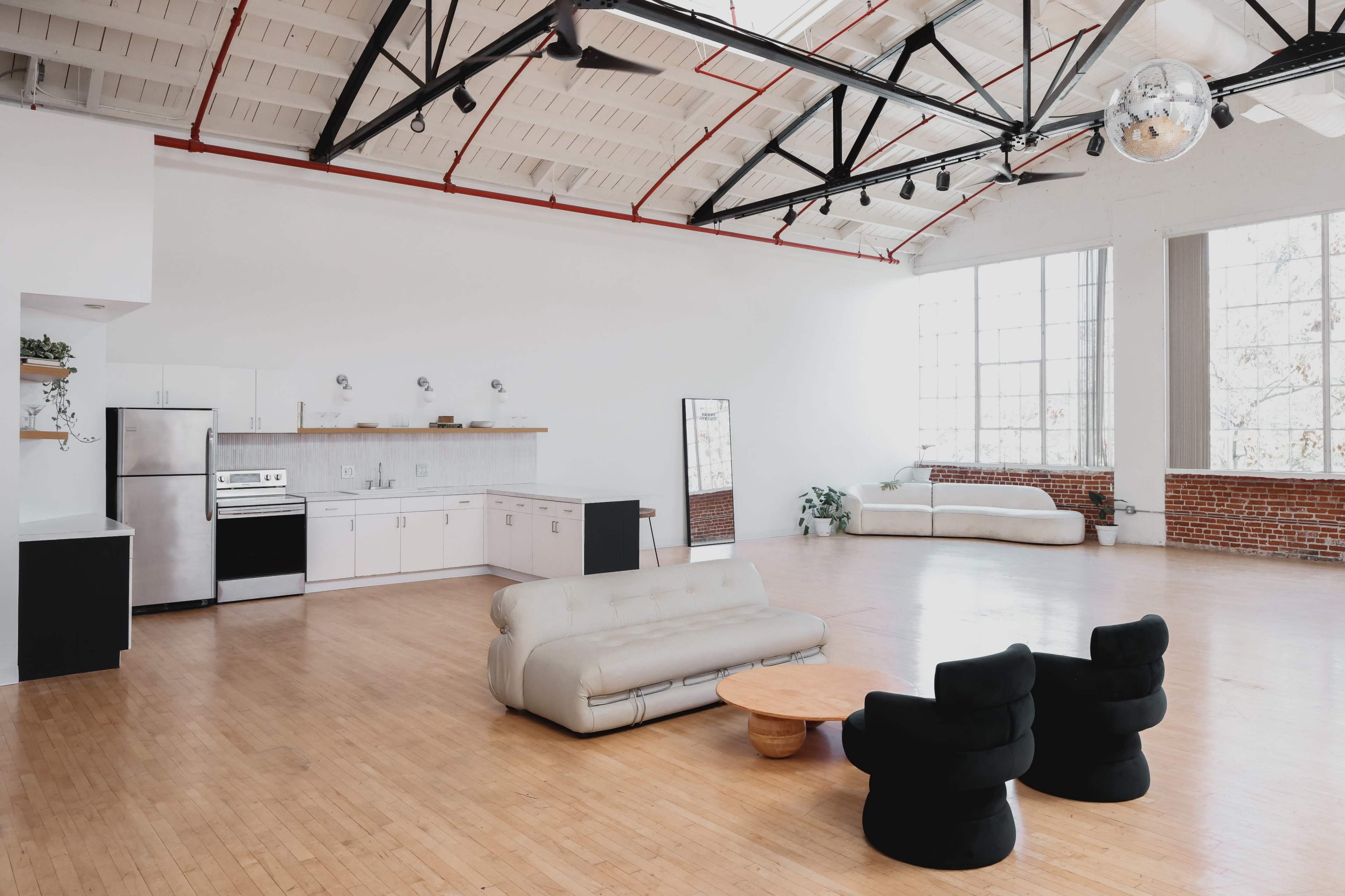 A spacious, modern loft with a kitchen and living area featuring a light-colored sofa, black armchairs, and large windows.