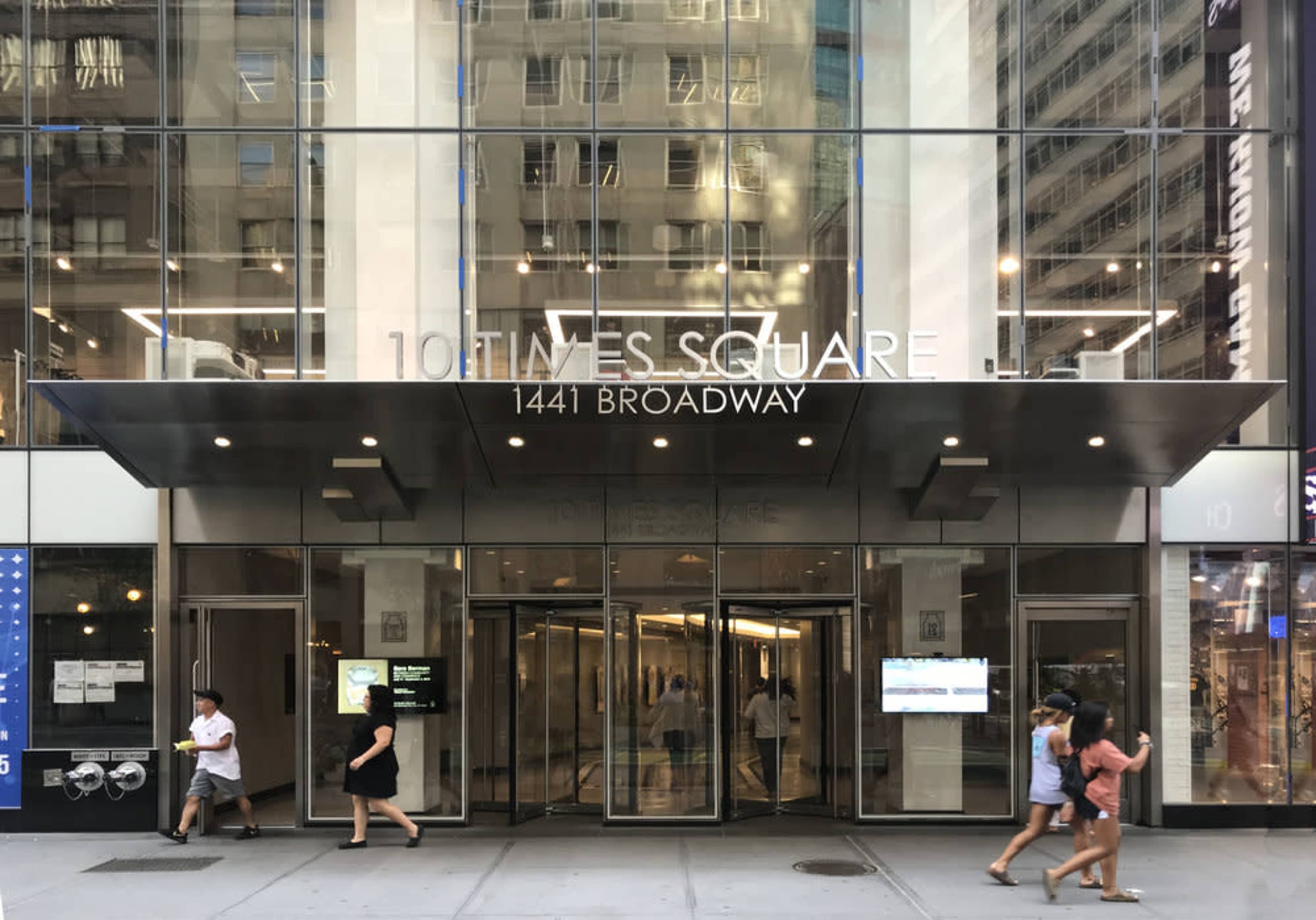The glass entrance of 10 Times Square at 1441 Broadway, with people walking past on the sidewalk.