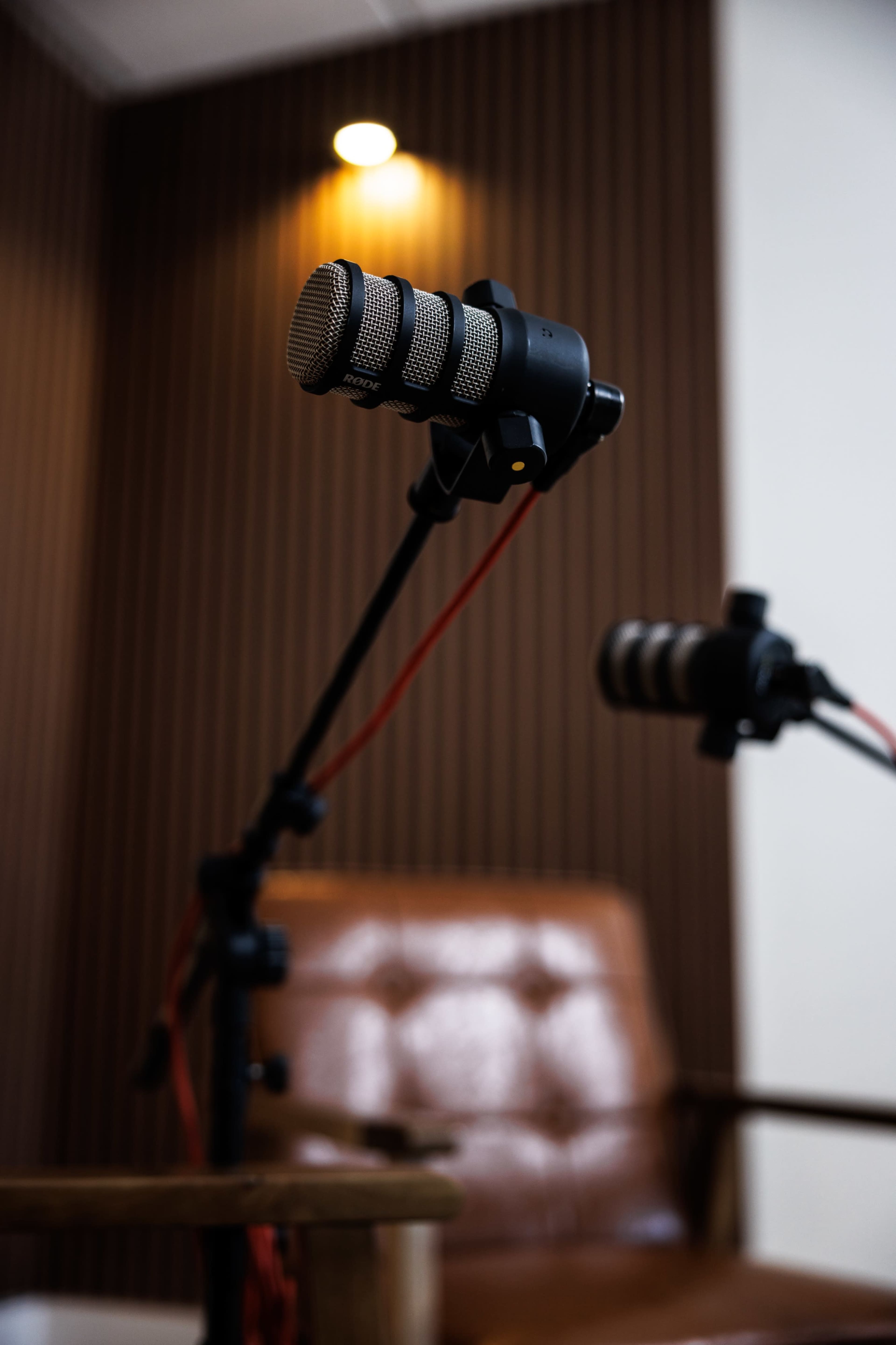 The image shows two microphones on adjustable stands positioned in front of a brown leather chair in a studio setting.