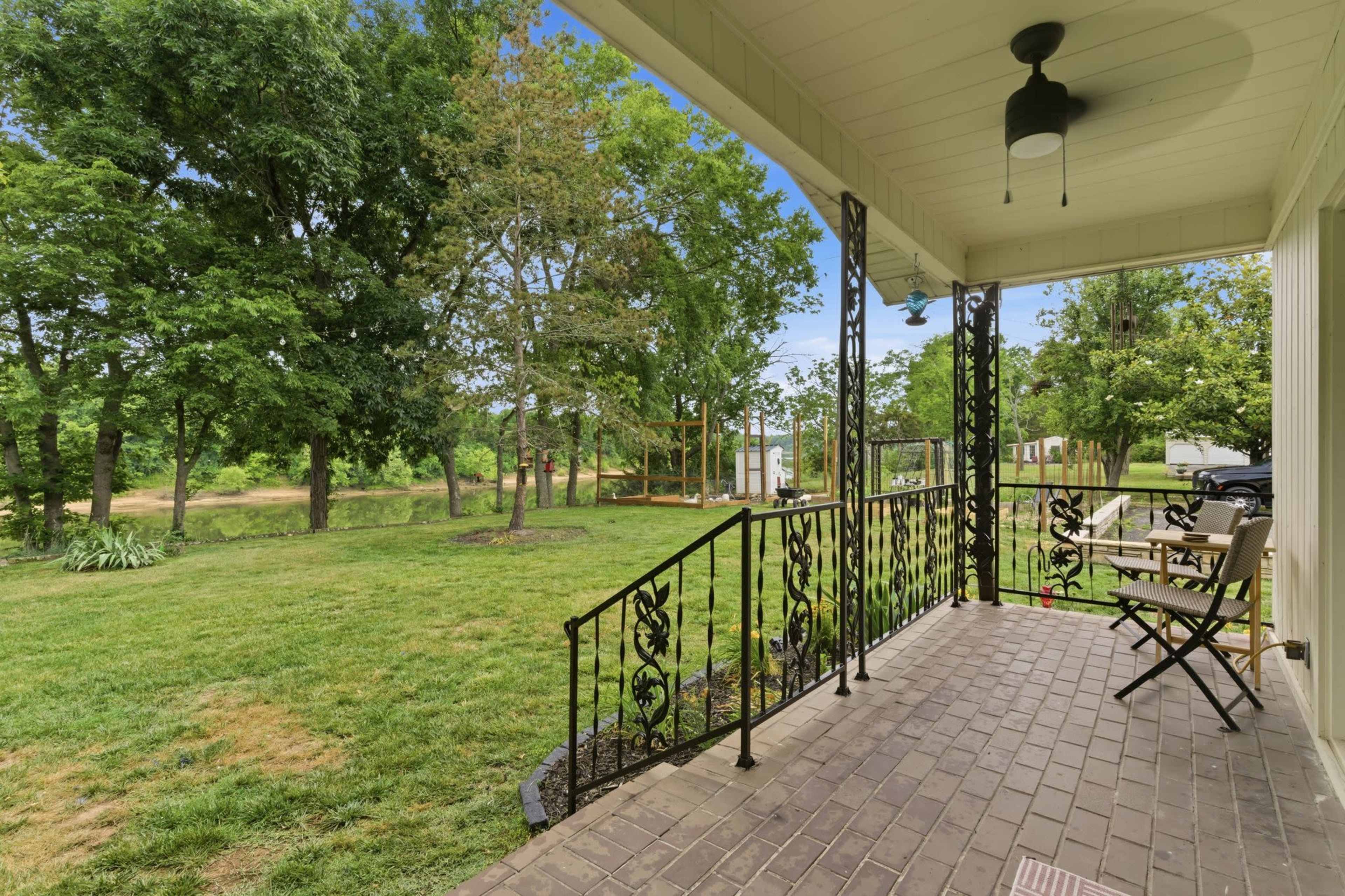 A porch features black metal railings and chairs, overlooking a grassy area with trees in the background.