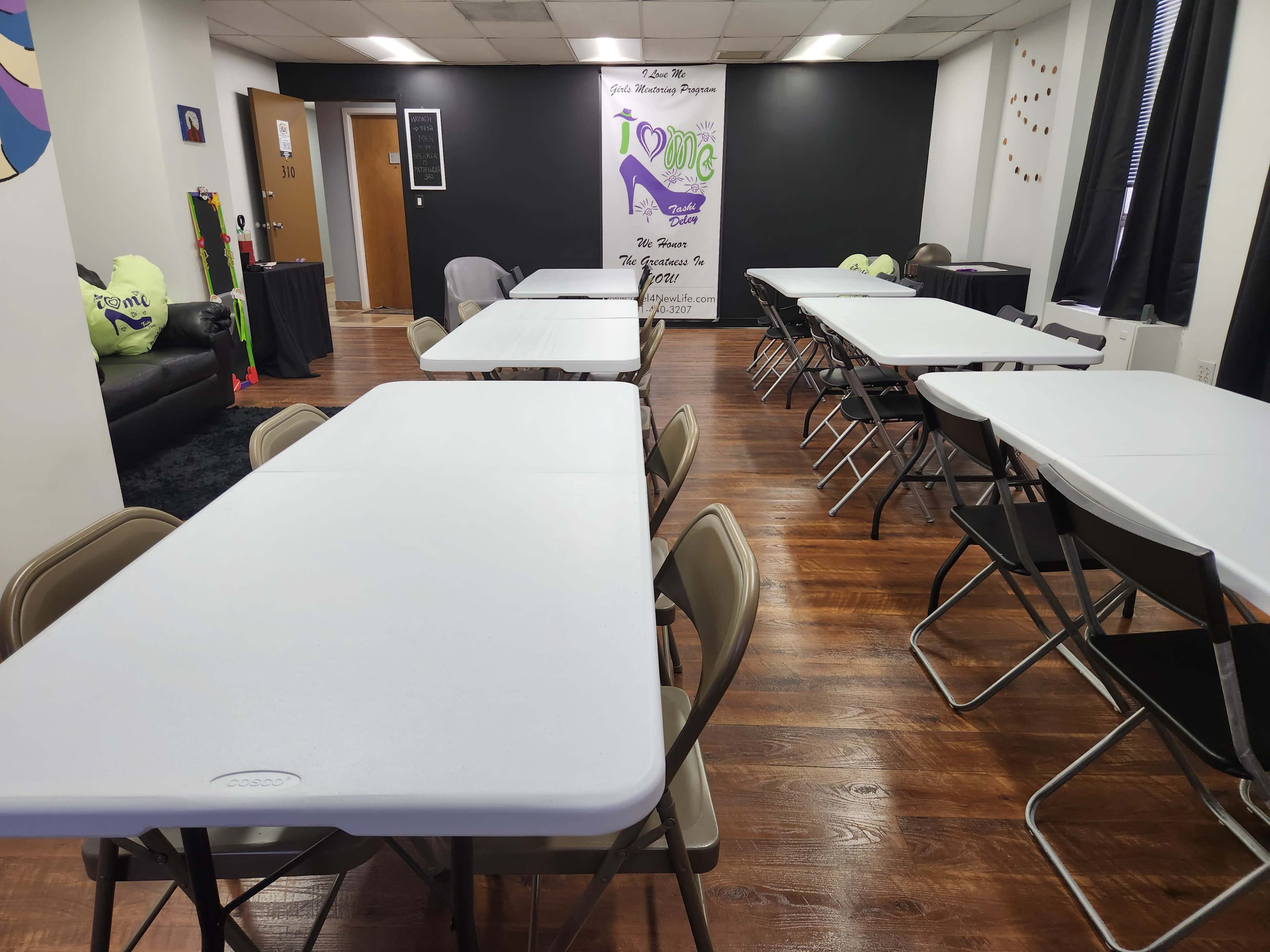 A large, well-lit room with multiple white folding tables arranged in rows, surrounded by folding chairs, and a black wall featuring a colorful poster.