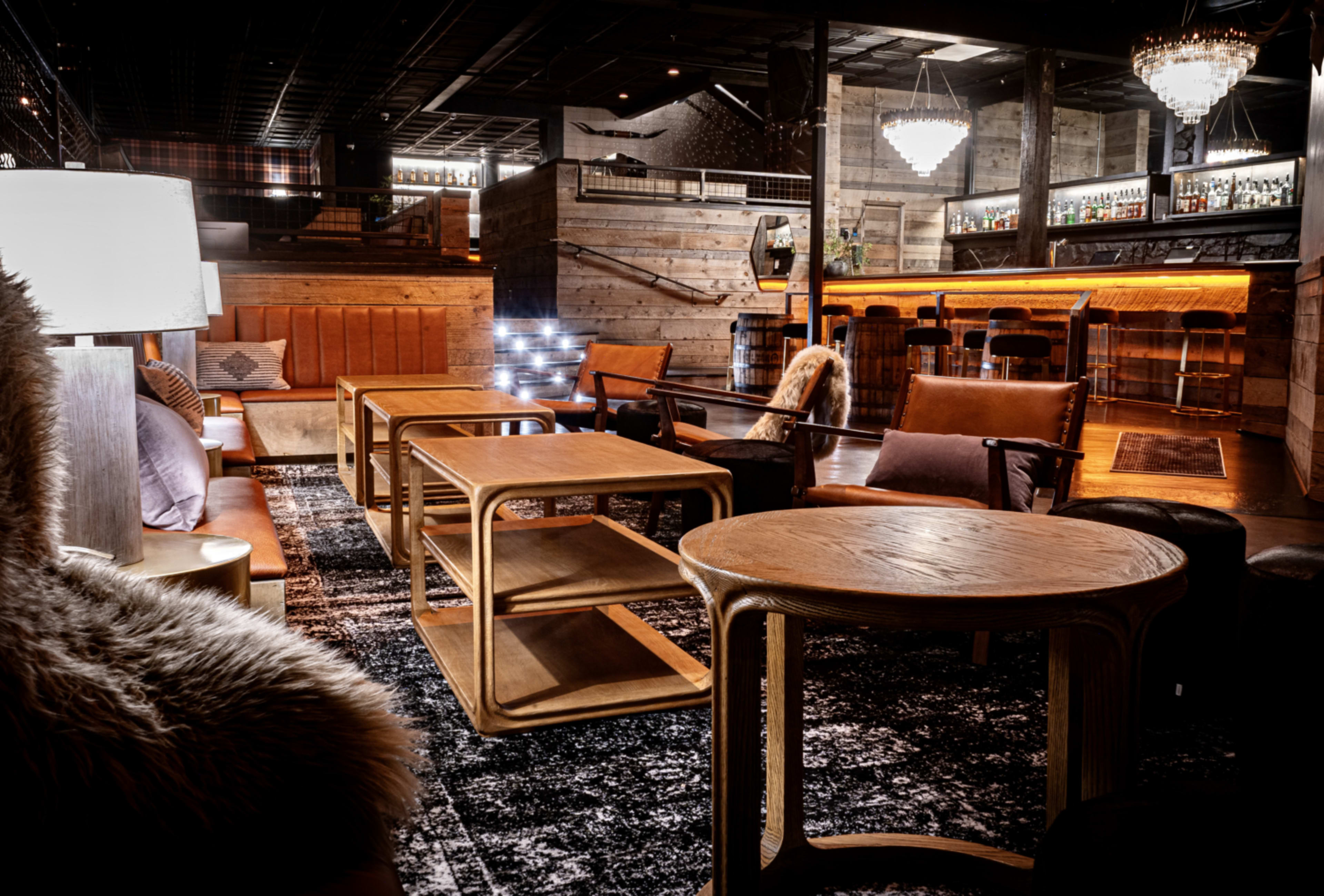 A stylish lounge area with wooden furniture, a patterned rug, and a well-lit bar in the background.