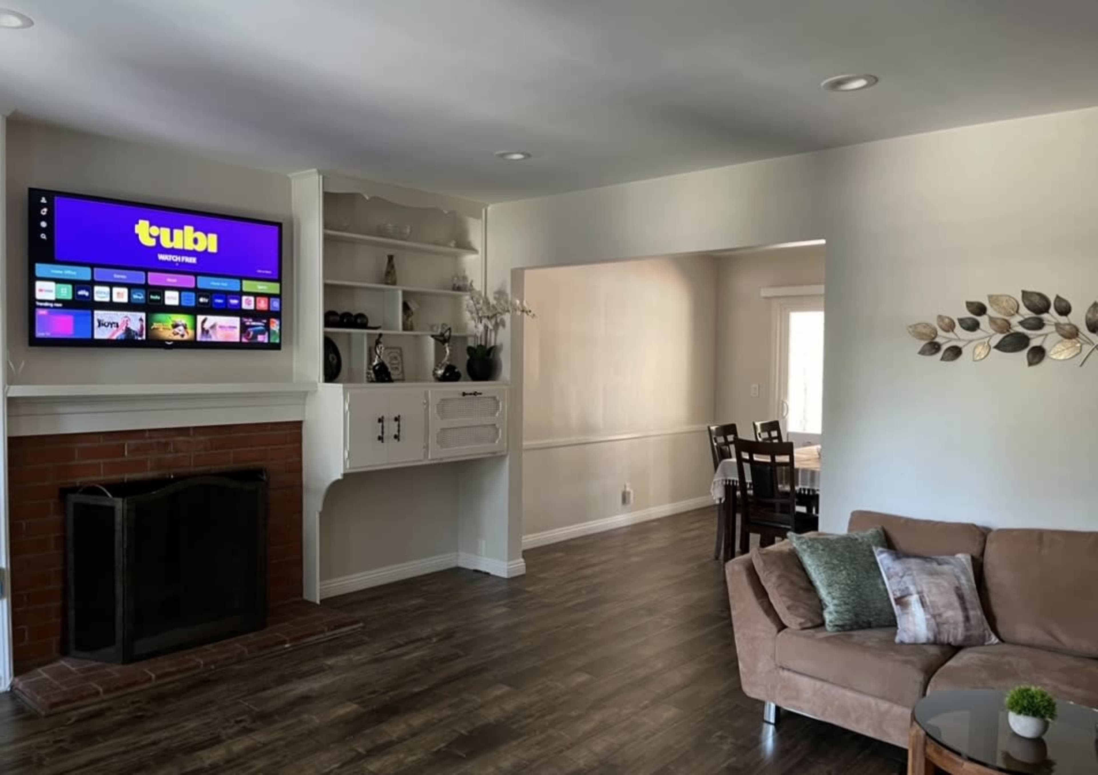 The image shows a living room with a mounted television above a brick fireplace, a beige sofa, and a dining area visible through an archway.