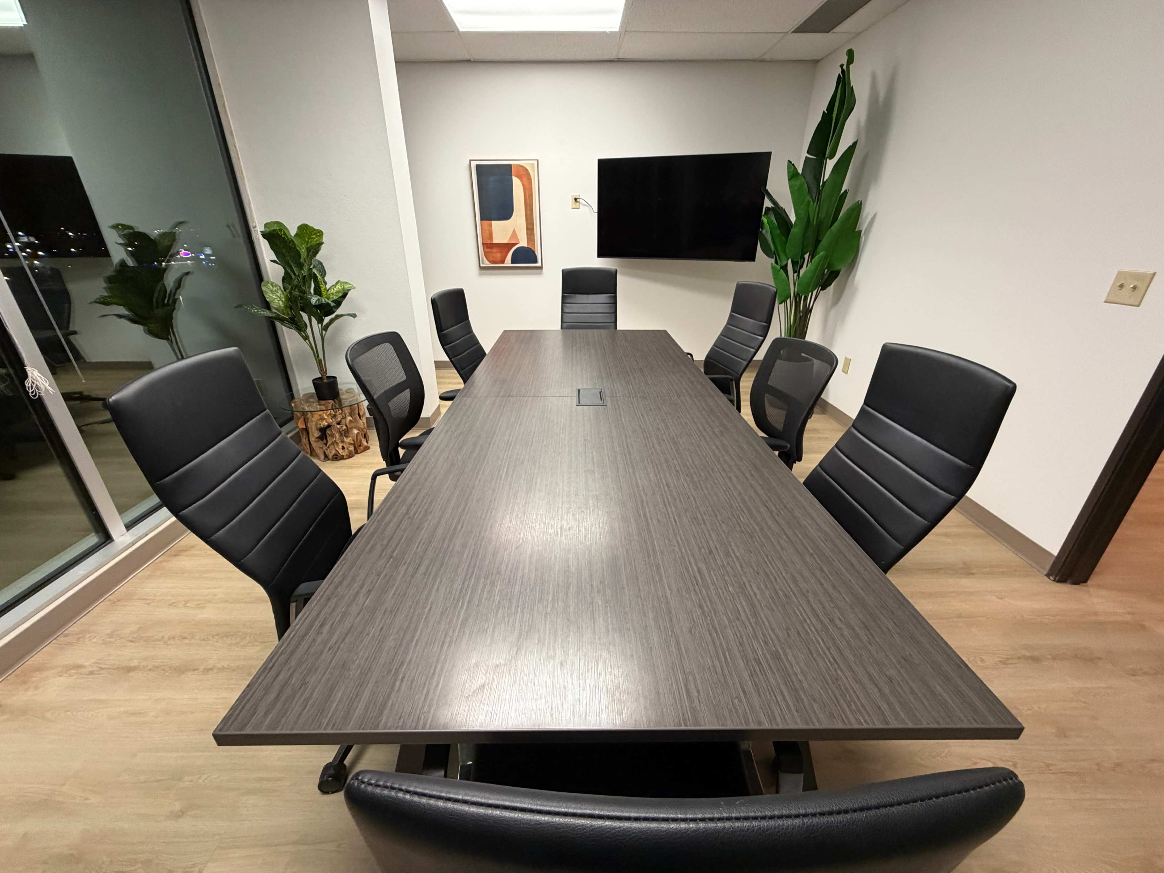 A long conference table surrounded by ergonomic chairs is situated in a modern meeting room with a large window and plants.