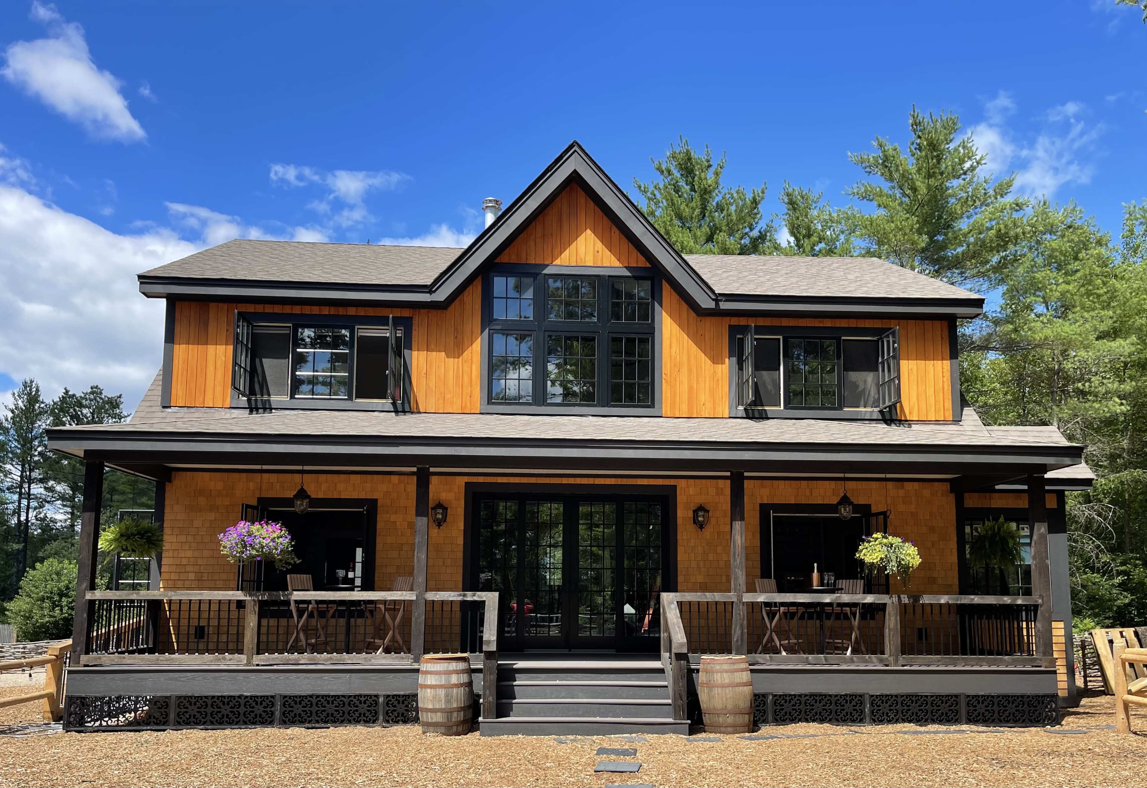 A two-story house with a wooden exterior, large windows, and a covered porch, surrounded by trees and gravel.