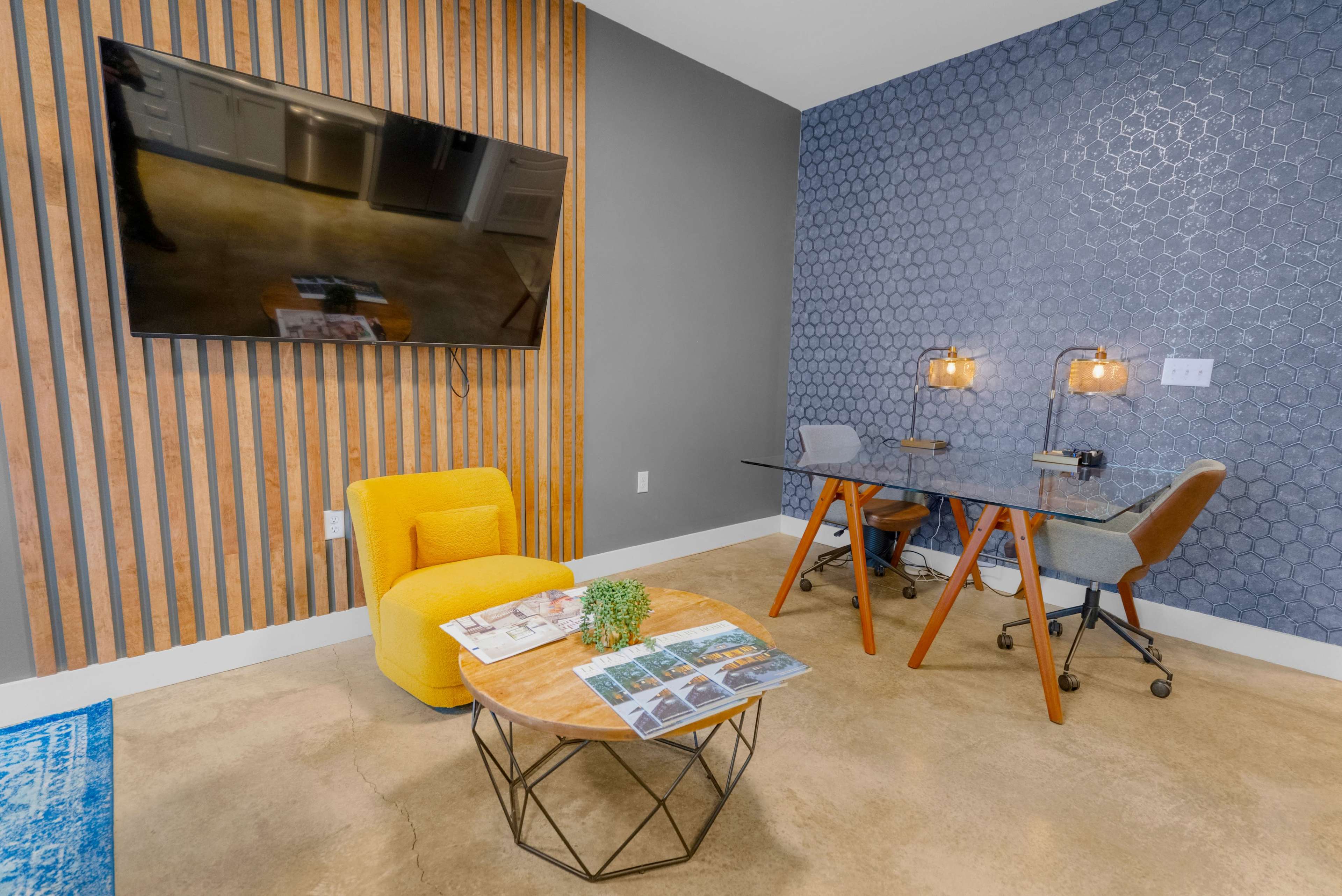 A modern office space featuring a yellow armchair, a coffee table with magazines, a glass desk with task lamps, and a television mounted on a wooden wall.