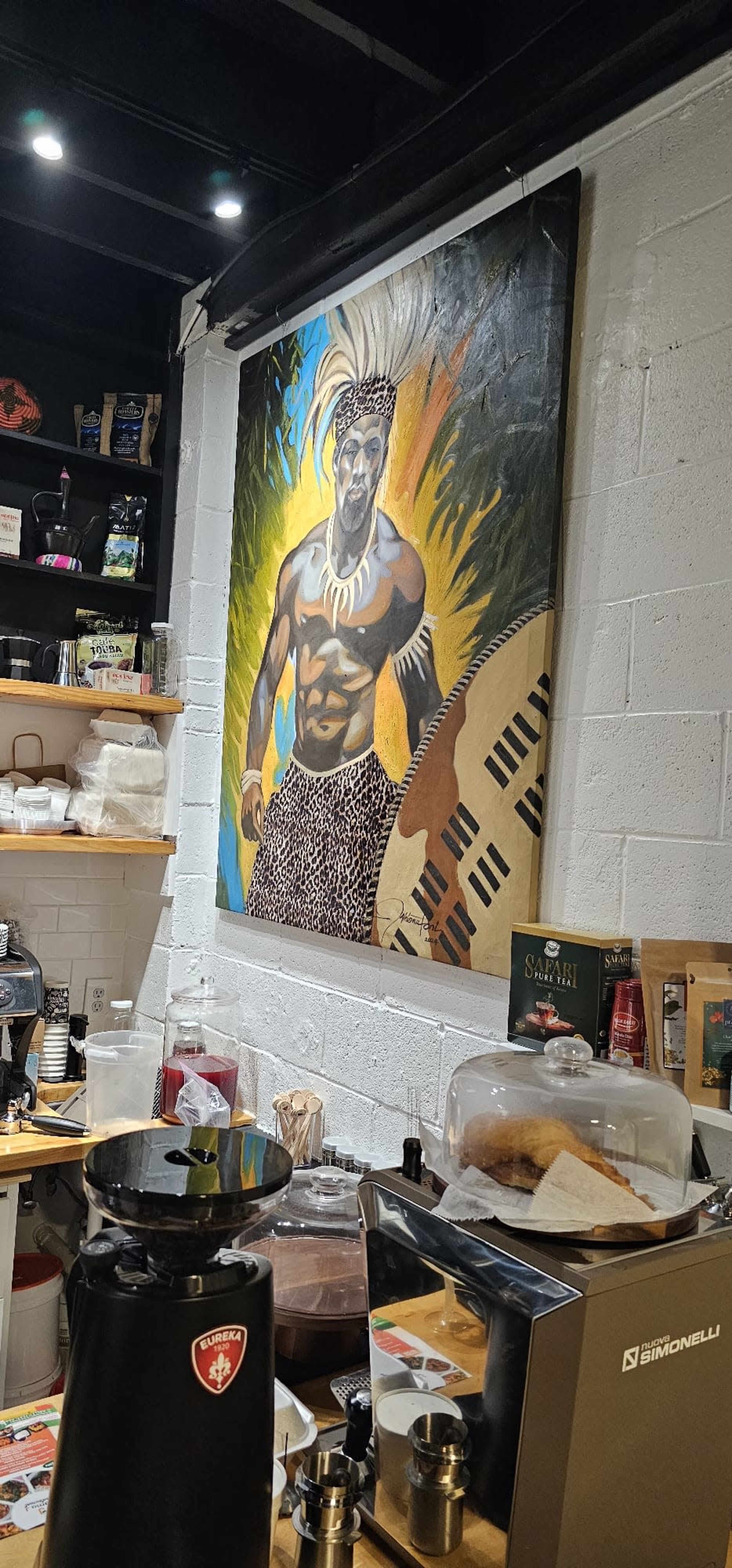 The image shows a coffee shop interior with a large, colorful painting of a man wearing traditional attire on the wall.