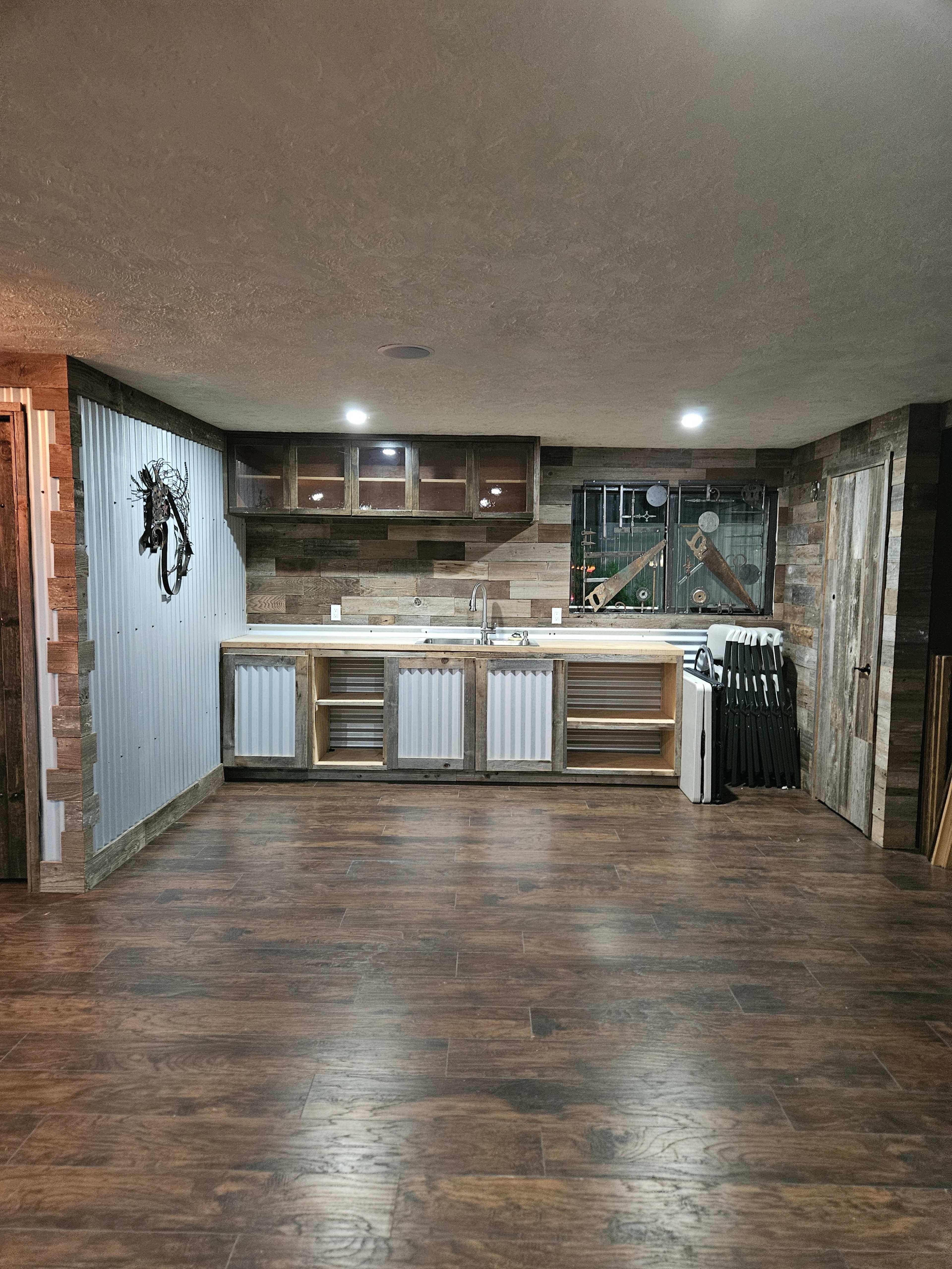 The image shows a spacious basement area featuring a modern kitchenette with wooden cabinetry and a countertop, as well as a wooden floor and various storage items against the walls.