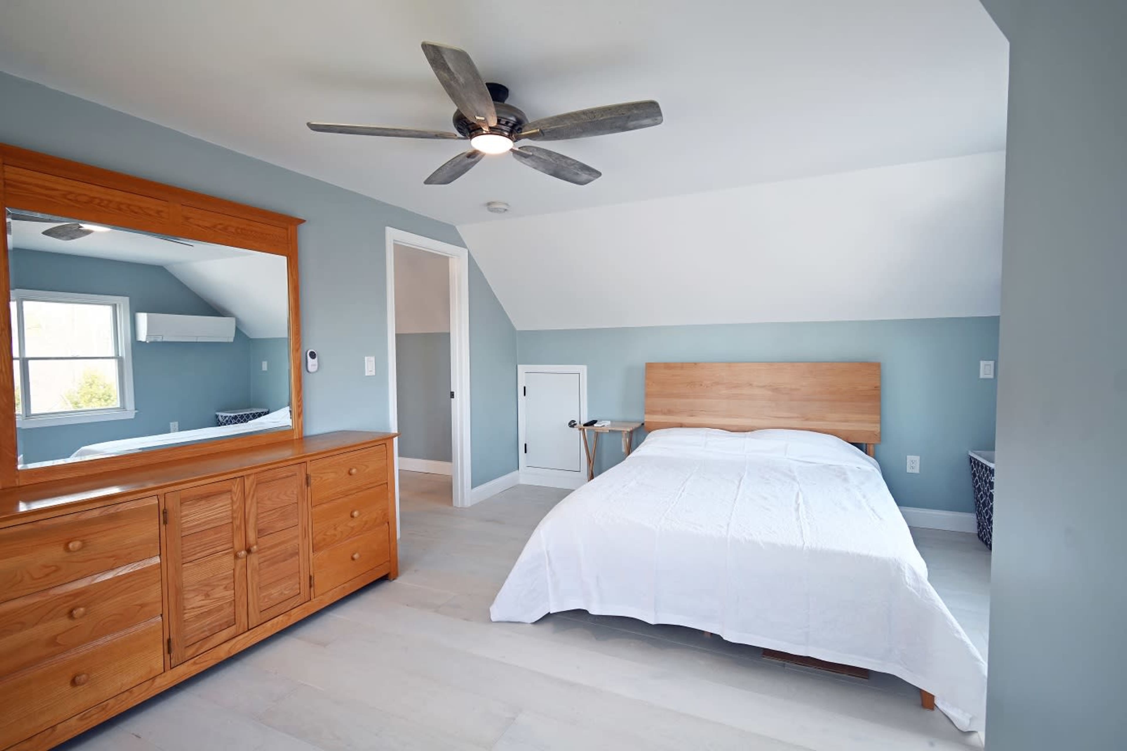 A simple bedroom featuring a bed with a white coverlet, a wooden dresser with a large mirror, and light blue walls.